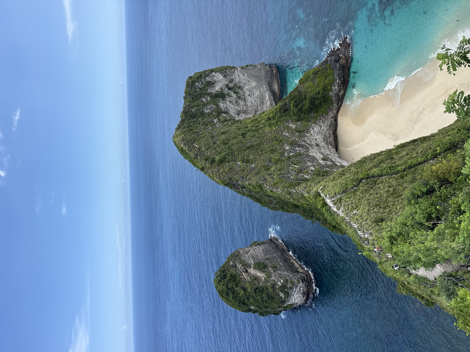 The image presents an aerial view of Nusa Penida's iconic Kelingking Beach, situated near Bunga Mekar, Indonesia.
A distinctive, T-Rex-shaped cliff dominates the frame. Its vegetation-covered spine descends steeply towards a secluded beach. The beach displays golden sand and turquoise waters. The ocean is a deep blue in the foreground.
A small, rocky island stands detached from the main cliff face. A visible pathway descends down the cliff, revealing the presence of people.