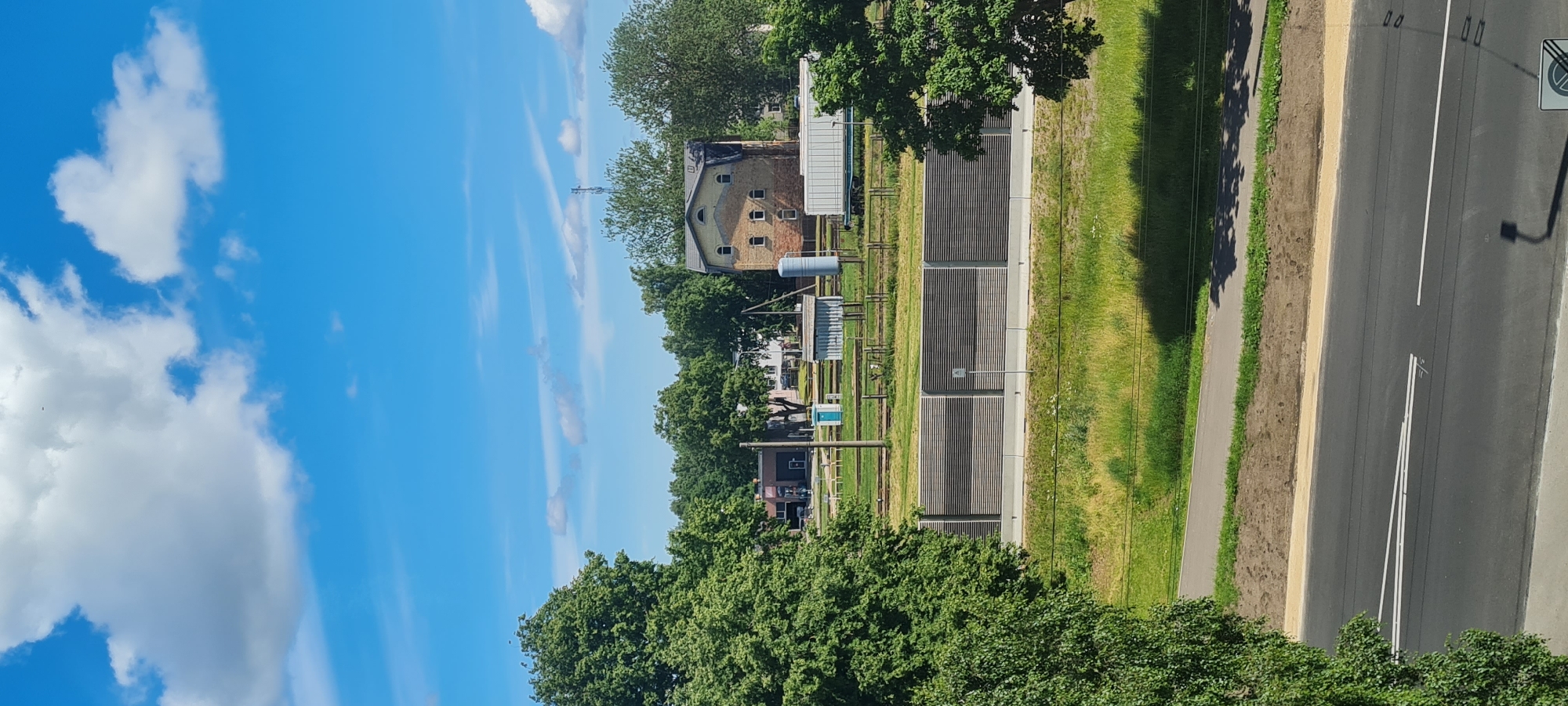 The image depicts a view of a partially urbanized landscape under a bright blue sky with scattered white clouds. In the foreground, a grey asphalt road with white lane markings is visible, suggesting a traffic or transit area. Beyond the road, a strip of green grass transitions into a grassy embankment and then a concrete barrier topped with a slatted noise reduction fence.
Further back, a line of trees with lush green foliage forms a backdrop. Behind the trees, several structures are visible. On the left, a dark building with a pitched roof and possibly a garage door is present. In the center, a tall, narrow, brick building with a pitched yellow roof and several windows is a prominent feature. Adjacent to this building are smaller structures, including what appears to be a water tower or large cylindrical tank and a shed-like building. A portable toilet is also situated in this area. The ground appears to be a mix of grass and gravel, with railway tracks visible in the distance. The presence of a tall communication tower further in the background indicates a developed area. The overall impression is a scene of mixed infrastructure and natural elements, possibly on the outskirts of a city.
This scene is likely located near the Jūrmalas gatve in Riga, Latvia, specifically in the vicinity of the "Bolderājas dambis" area, known for its industrial and transit infrastructure. The noise reduction fence along the road is a common feature along major transit routes in Riga to mitigate sound pollution. The juxtaposition of the road, fence, and distant buildings suggests a location that has undergone recent development or infrastructure upgrades.