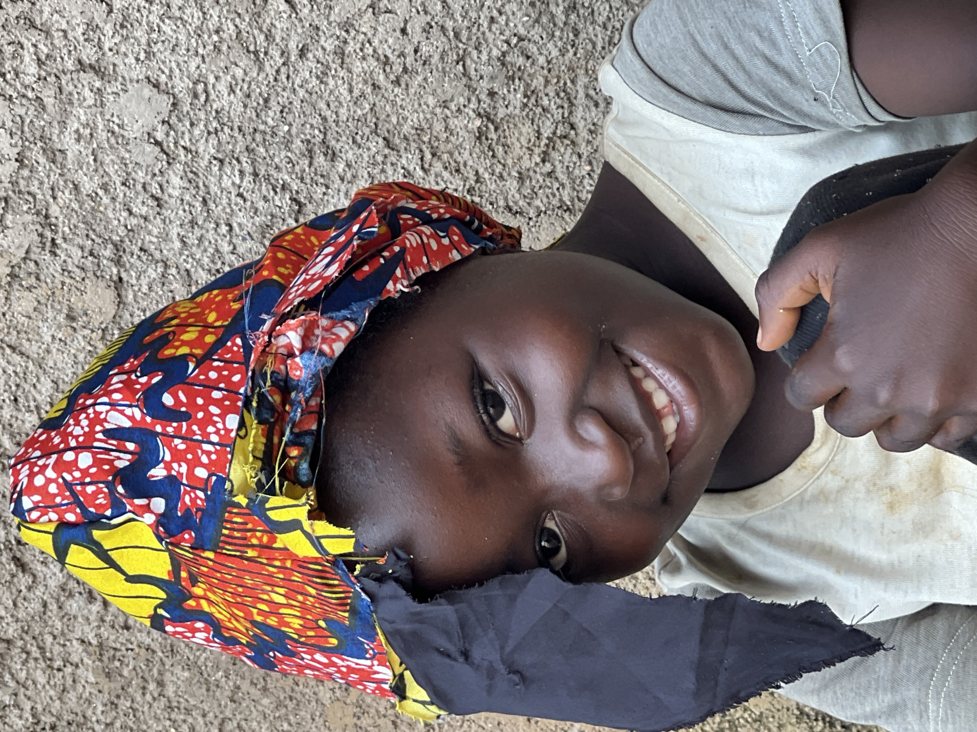A close-up, waist-up shot shows a young Black girl with a wide, toothy smile and dark eyes looking directly at the camera. She is wearing a colorful headwrap with vibrant red, yellow, and blue patterns, and a simple beige t-shirt with gray sleeves. Her left hand is holding a piece of dark fabric that is partially covering her face. The background is a textured, pale gray wall. The image was captured in Jos, Nigeria, suggesting an outdoor setting in a city.