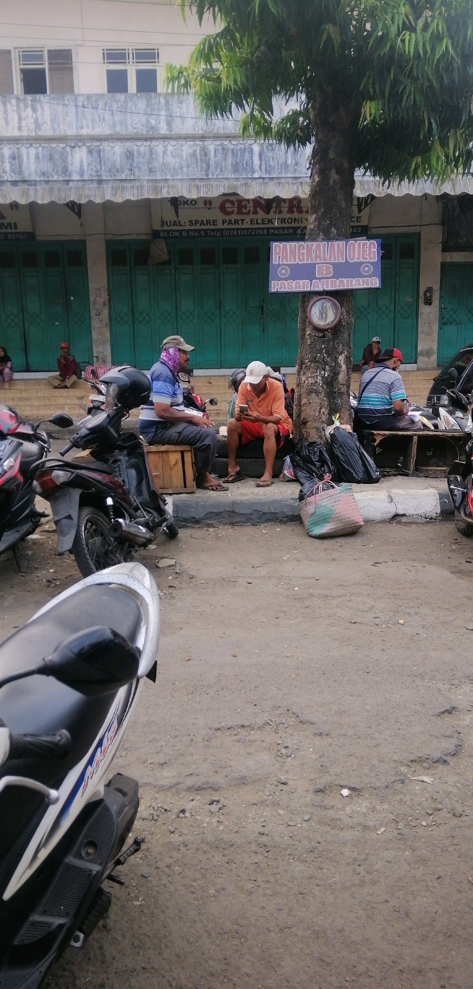 A street scene in Ajibarang, Indonesia features a group of men gathered near a tree. The men are seated on wooden crates and tires, with several motorcycles parked nearby. One man is looking at his phone, while others appear to be conversing or resting. In the background, a building with green shutters and a sign that reads "PANGKALAN OJEG B PASAR AJIBARANG" (OJEG Base B Ajibarang Market) is visible. The ground is a mix of dirt and asphalt, with some debris scattered around. A woven bag and black trash bags are placed near the men. The overall atmosphere suggests a typical day at a local transportation hub in a small Indonesian town.