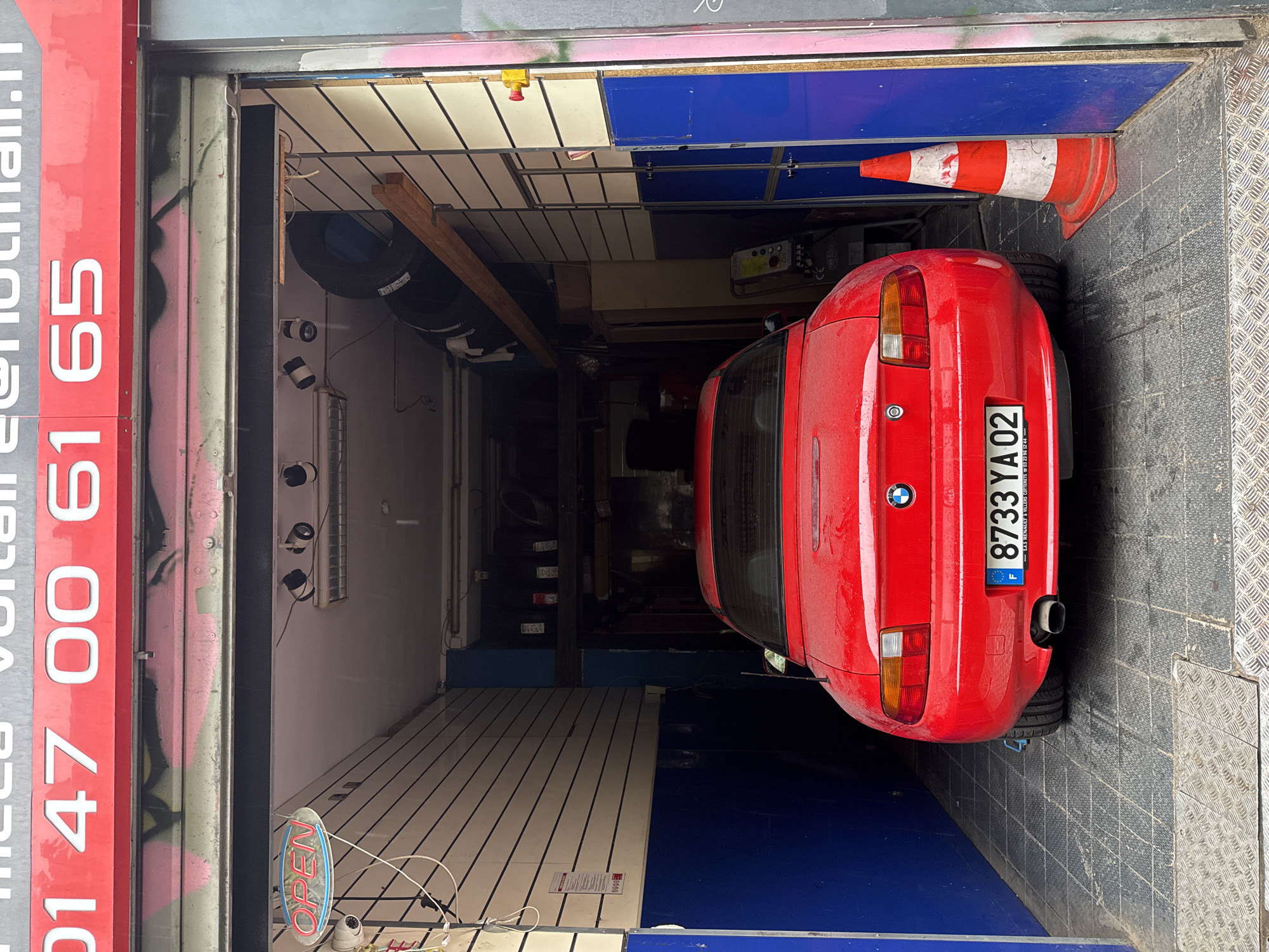 The image shows a red BMW Z3 parked inside a garage in Paris, France. The license plate is "8733 YA 02," which appears to be a French registration. The garage door is partially open, revealing the interior with various items, including tires and equipment. There is a red and white traffic cone near the car. An "OPEN" sign hangs near the entrance. The exterior of the building features red signage with white text, "01 47 00 61 65." The address appears to be associated with "SAS Renault & Villers Cotterets 03 23 96 12 44."