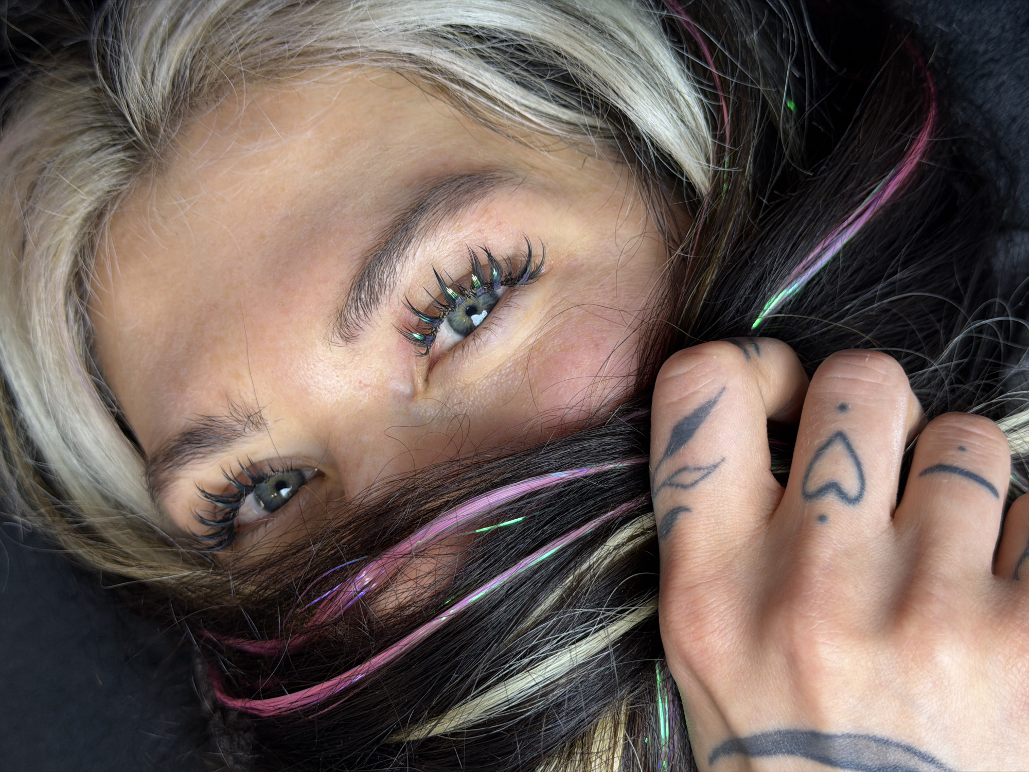 A close-up, high-angle shot depicts a person's face obscured by their hair, with focus on the eyes and the right hand. The hair is multi-colored, featuring blonde, black, and strands with pink and green iridescent tinsel. The person's light-colored eyes are partially visible amidst the hair. They have dark eyelashes, enhanced with reflective green details. The right hand, adorned with several small black tattoos including a heart, a line, and a leaf design, is seen holding or pulling the hair away from the face. The skin tone is fair. The background is dark and out of focus. The location in the city of Budva, Montenegro cannot be determined from this image.
