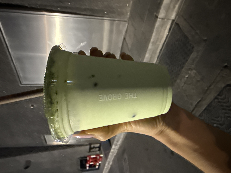 A hand holds a plastic cup of a pale green drink. "THE GROVE" is embossed vertically on the cup. The lid is translucent. The drink appears to be a matcha beverage with dark flecks throughout. The background is a mix of stainless steel and gray textured surfaces, possibly in a commercial kitchen or food service area. A small, red and black object is visible in the lower left corner of the frame.
