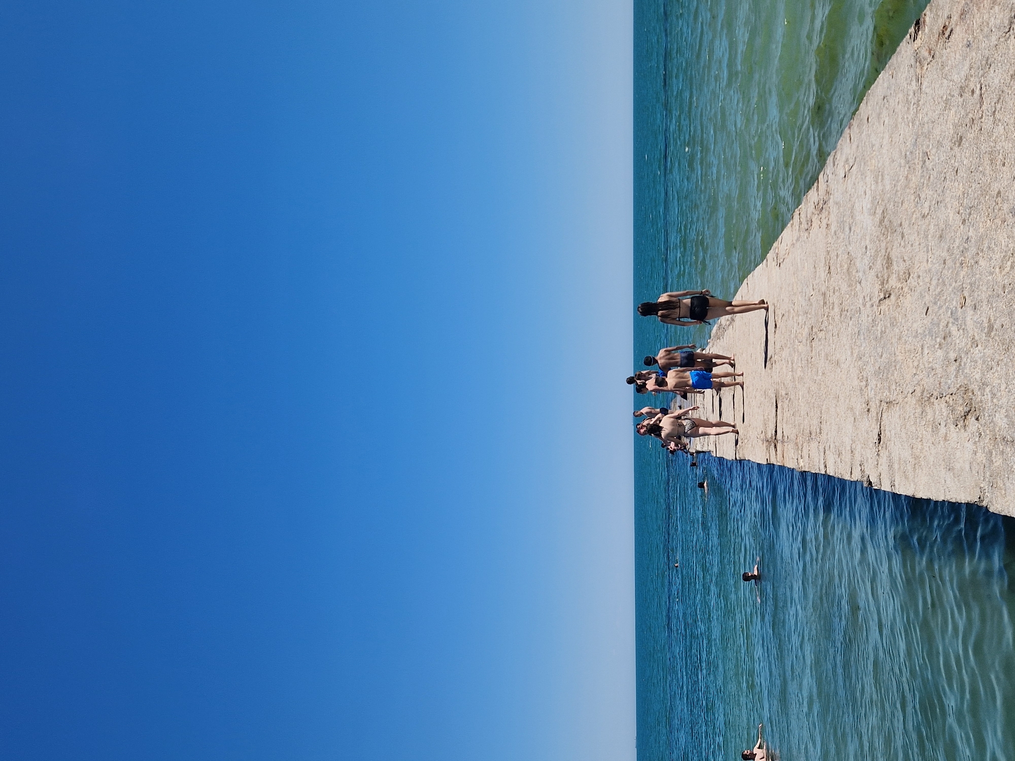This is a vertical, eye-level, full-shot photograph of people on a concrete pier next to the ocean. The sky takes up the left two-thirds of the image and is a clear, bright blue, transitioning to a lighter hue near the horizon. The right third is the ocean, a vibrant turquoise, with small waves visible.
A group of people are scattered across the pier. In the foreground, closer to the water's edge, several individuals are standing or sitting. One person in a dark bikini is facing away from the camera, with their back to the sea, and their legs extended. Further up the pier, a cluster of people are also standing, some facing away, others looking down at the water. There are more people in the water, appearing to swim or float. One person is visible with their head above the water.
The scene appears to be set on a bright, sunny day, likely during the summer, given the attire of the people and the clear weather. The concrete pier looks weathered and solid. The overall mood is relaxed and summery, with people enjoying the water and the pier. There is no visible text in the image.