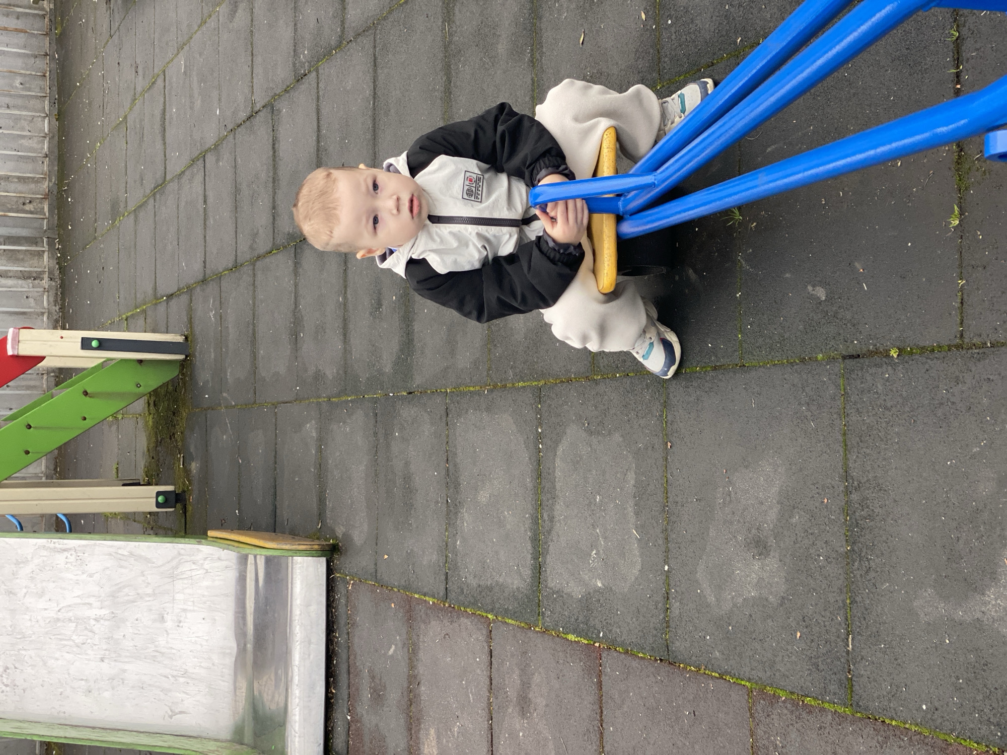 A young child sits on a blue and yellow playground seesaw. The playground surface is composed of interlocking grey tiles with some green moss in the cracks. A metal and green structure with a ladder is visible in the lower left corner. The setting appears to be in Sochi, Russia, though this cannot be confirmed from the image alone. The child is wearing a white and black jacket, light-colored pants, and sneakers.