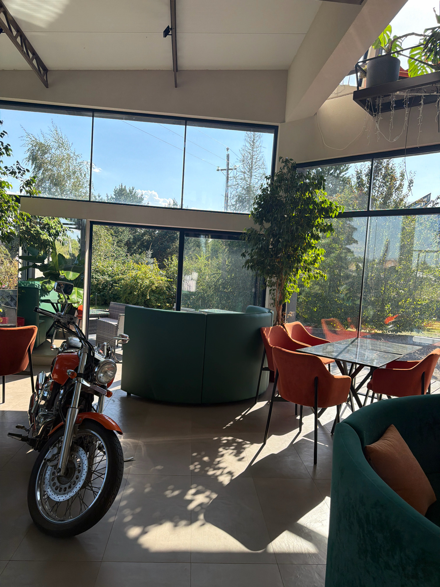 The image depicts the interior of a contemporary establishment, likely a cafe or restaurant, in Malaya Tsarevschina, Russia. The space is characterized by large, floor-to-ceiling windows that offer a view of lush greenery and a bright sky, suggesting an outdoor seating area or garden. Inside, the furniture is modern and stylish. A vibrant orange motorcycle is parked prominently in the foreground, positioned to the left of the frame. The motorcycle appears to be a cruiser-style bike, with chrome accents and a single headlight.
Seating arrangements include plush, dark green sofas and armchairs, complemented by orange upholstered chairs surrounding a dark, reflective table. A tall, artificial tree stands near the windows, adding a touch of nature to the indoor environment. The floor is tiled, and the sunlight streaming through the windows casts dynamic shadows from the foliage and structural elements onto the floor. Overhead, exposed beams and lighting fixtures contribute to the modern aesthetic. The overall impression is one of a well-lit, aesthetically pleasing space that blends indoor comfort with outdoor views. There are no people visible in the image.