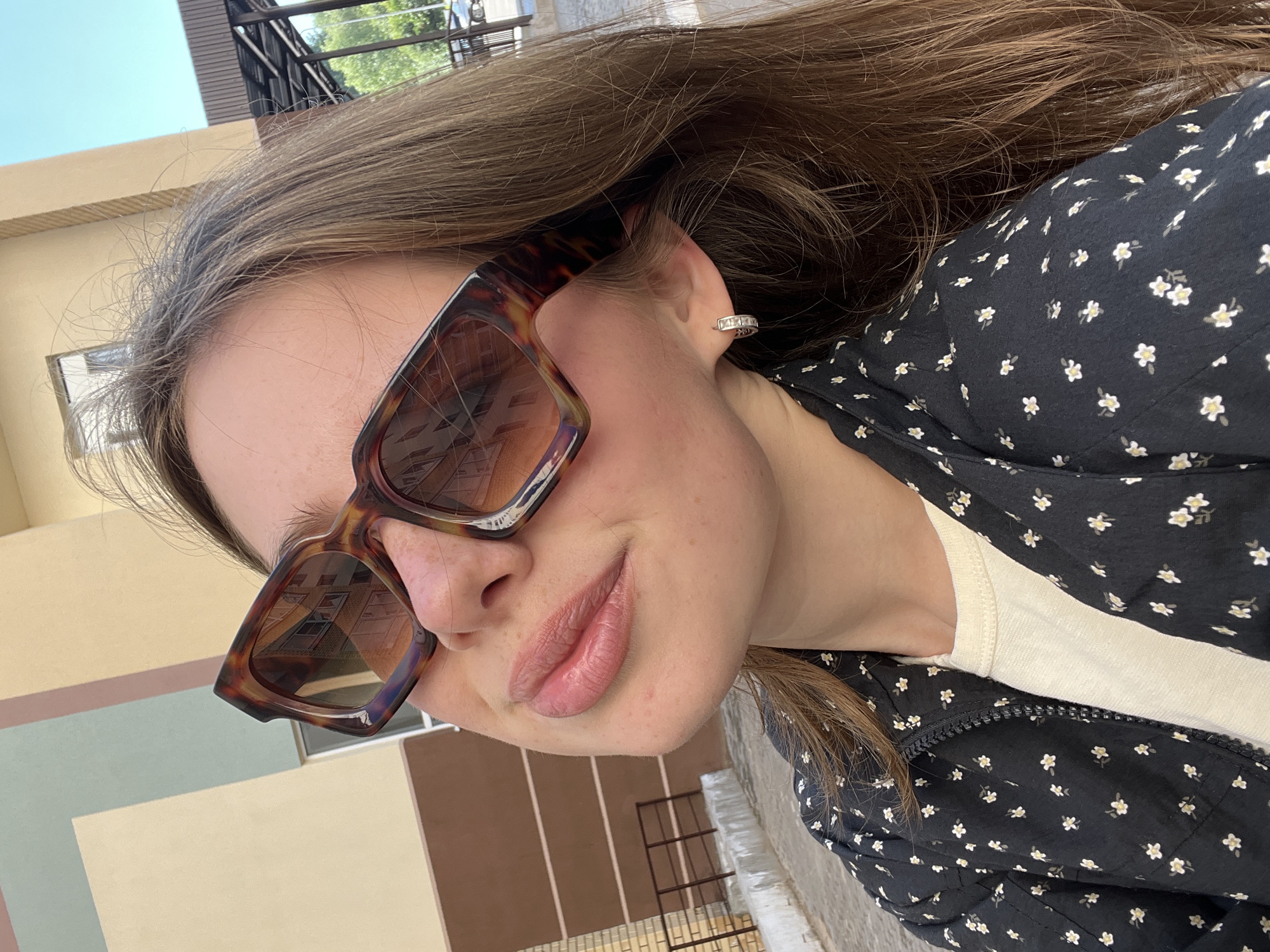 A young woman with long brown hair and fair skin is photographed outdoors in a city. She is wearing large, tortoise-shell patterned sunglasses, a white t-shirt, and a black jacket with a small white floral print. She has a silver hoop earring with small stones in her left ear. Her lips are slightly parted and appear to have a light pink lip gloss. The background shows a building with beige and brown sections, a metal fence, and a blue sky. The reflection in her sunglasses shows a building with windows. The location is identified as Chaikovsky, Russia.