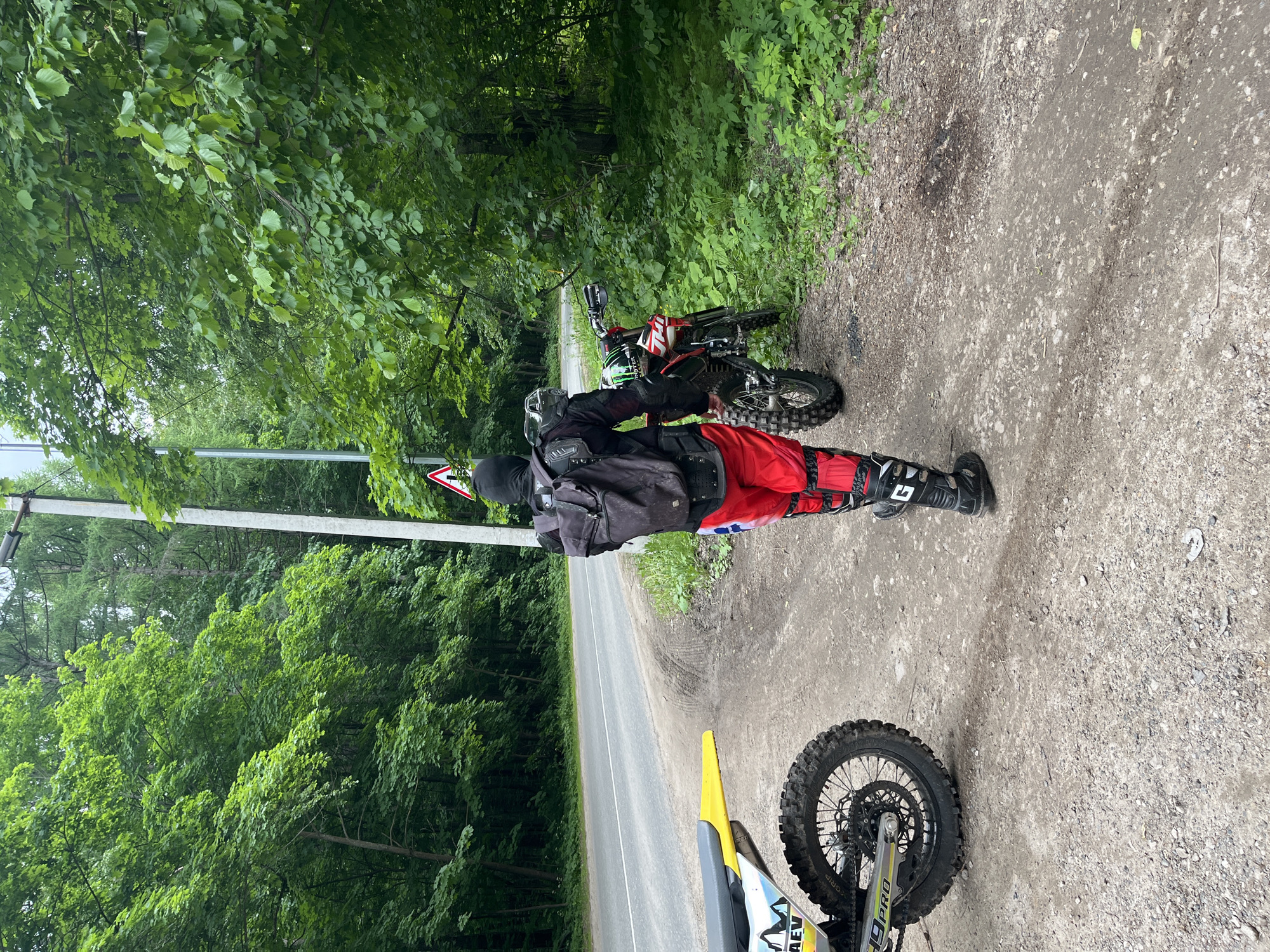 The scene is located in Ореховка, Russia, depicting a dirt shoulder adjacent to an asphalt road bordered by dense green deciduous trees under an overcast sky. One person in full motocross riding gear is positioned on the dirt shoulder, facing towards a red and white dirt bike. The individual wears a black protective jacket or vest, a black balaclava, red motocross pants with white and black accents, and black motocross boots with a visible "G" logo on the shin guard. A black, visibly dirty backpack is worn over the protective jacket. A green helmet, featuring a Monster Energy logo, rests on the handlebars of the red and white dirt bike. A second dirt bike, partially visible on the lower left, has a yellow rear fender, a grey seat, and a white side panel with "AEV" and "19 PRO" graphics. Both bikes are equipped with knobby off-road tires. The asphalt road to the left exhibits a faded double solid yellow centerline. A grey utility pole with a street lamp fixture and a red triangular warning sign is positioned on the right side of the image. The ground consists of light-colored dirt and gravel, with visible tire tracks.
