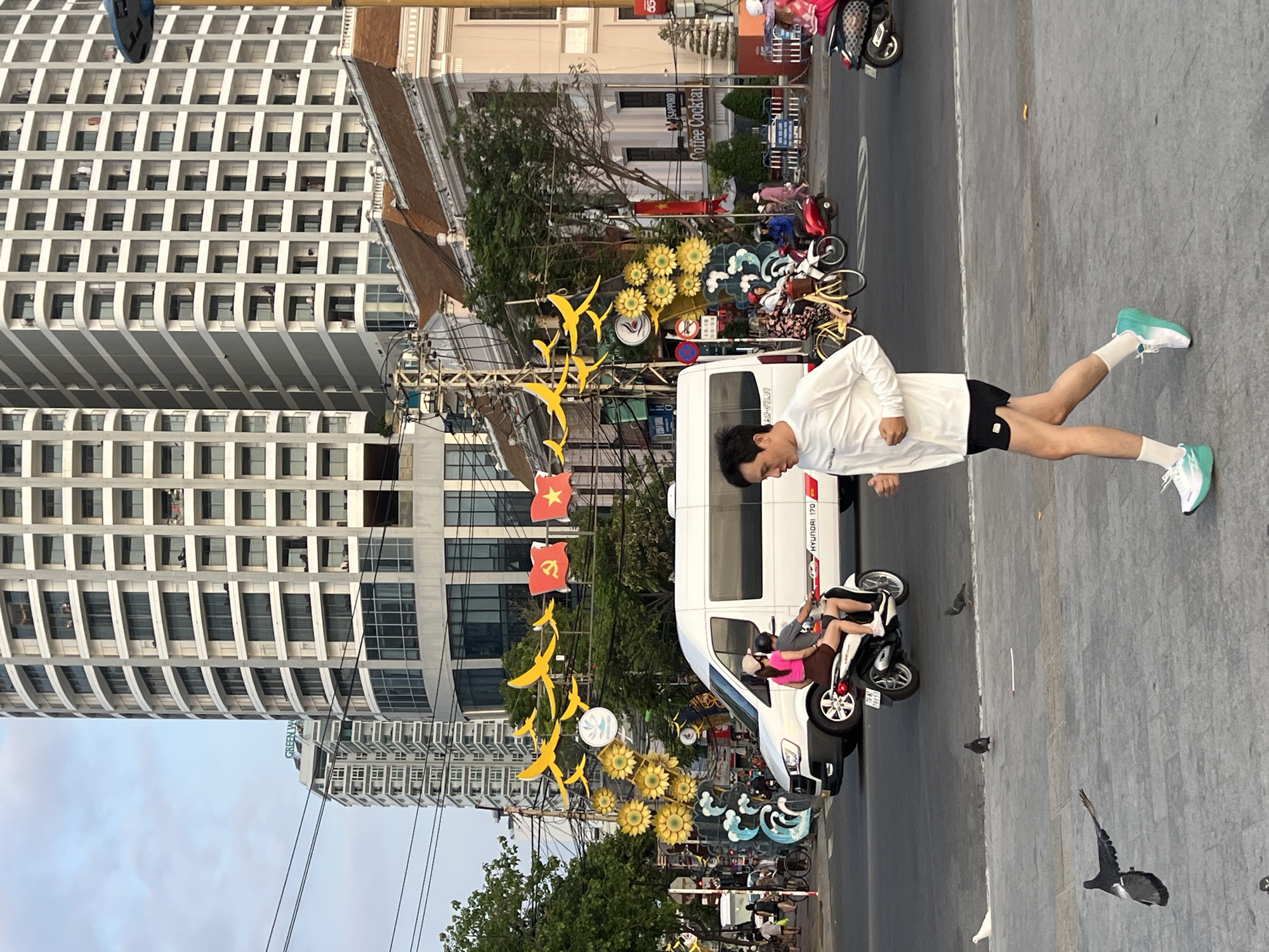 A horizontally oriented, outdoor photograph captures a street scene in Nha Trang, Vietnam, with a man jogging in the foreground.
The man, wearing a long-sleeved white shirt, black shorts, white socks, and turquoise sneakers, runs towards the right. Behind him, two pigeons are visible on the pavement, one in mid-flight.
A white van is parked on the street. Two individuals are riding a motorcycle next to the van. The street is filled with motorbikes and pedestrians. Buildings are visible in the background, including a tall, modern building with a grid-like facade, and other structures with pitched roofs.
Decorations, including yellow bird figures and sunflower decorations, are strung across the street along with flags, adding color to the scene. The sky is overcast.