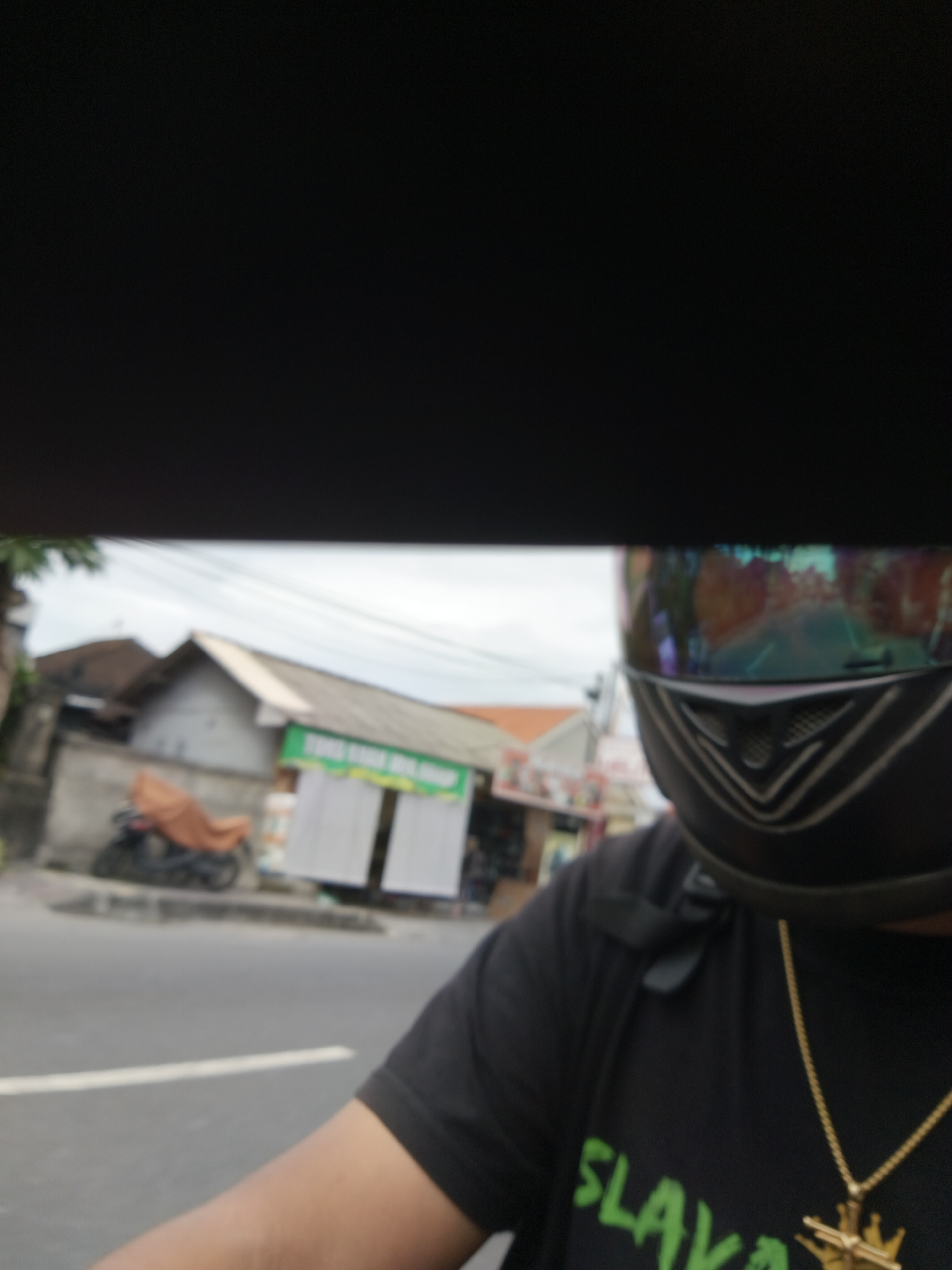 The image shows a first-person perspective of a person wearing a black t-shirt with a green graphic and a gold chain, as well as a black motorcycle helmet with a reflective visor. The person appears to be riding a motorcycle, as evidenced by the road and surrounding environment. In the background, there are buildings, including a structure with a green sign that is partially visible. A parked motorcycle covered with an orange tarp is also visible. The scene suggests an urban or suburban setting in Denpasar, Indonesia. The sky is overcast.