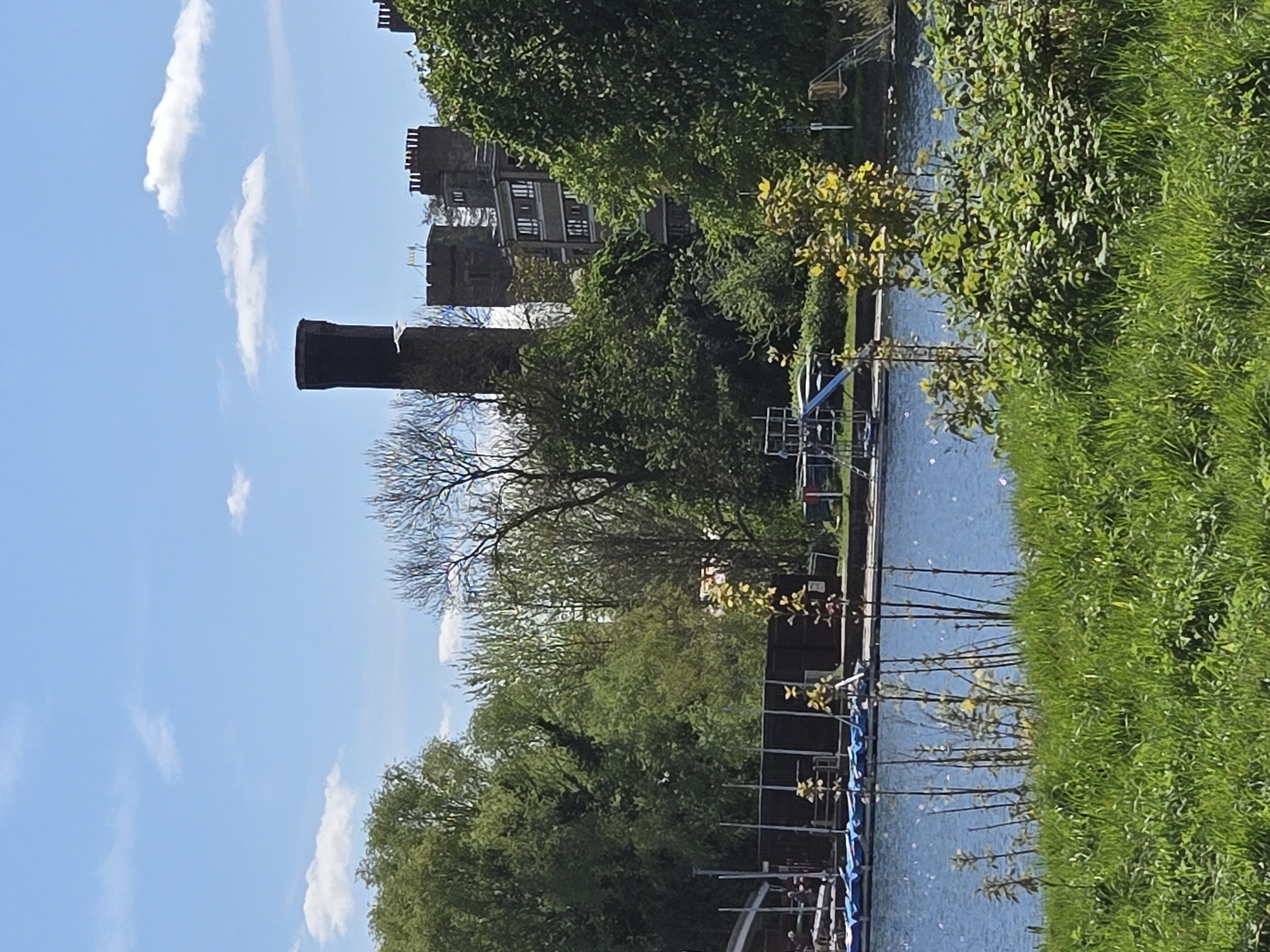 The image depicts a scene in London, United Kingdom, featuring the River Thames. In the foreground, lush green grass lines the riverbank. The river's surface is slightly rippled, reflecting the sky and surrounding foliage. Several metal frameworks or supports are visible extending into the water. In the midground, trees and bushes form a dense green backdrop. A tall, dark brick chimney stands prominently against the blue sky, adjacent to a building resembling a castle with turrets. Scattered clouds dot the clear, blue sky above. The general atmosphere suggests a sunny day in a park or recreational area along the Thames.