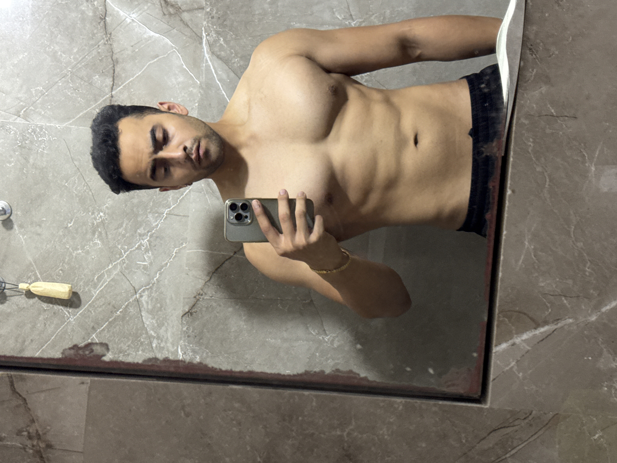 A shirtless man takes a selfie in a bathroom mirror. He has dark hair and a short beard, and he's wearing black shorts. He holds a silver smartphone in his right hand. The bathroom features gray marble walls and flooring. There is a small cleaning implement, a brush, stuck to the wall on the left. The city of Kolkata, India is not confirmed and cannot be included as part of the description.