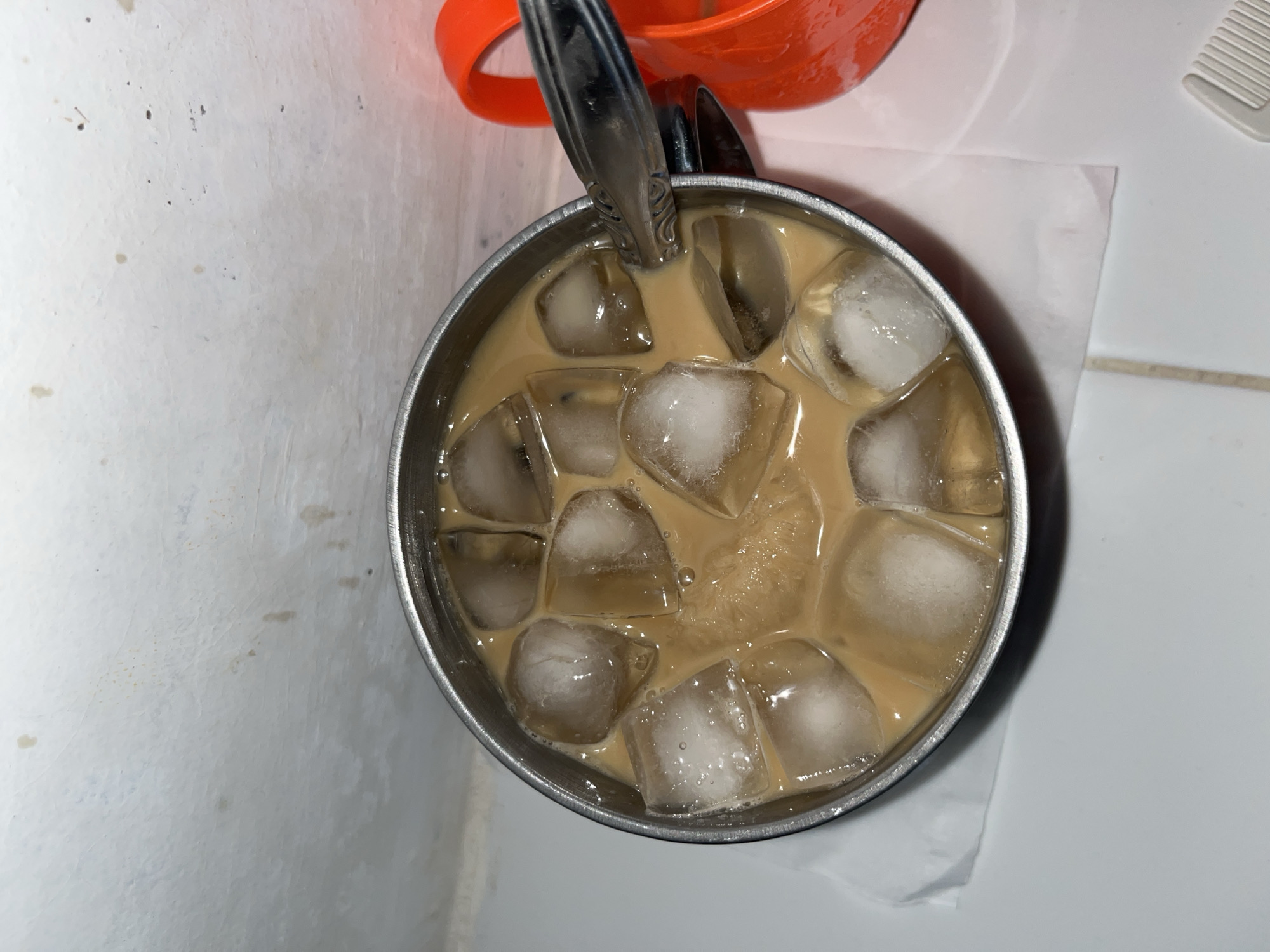 A close-up, top-down shot shows a metal cup filled with iced coffee. The coffee is a light brown color with frothy bubbles, and it is packed with ice cubes. A silver spoon is resting in the cup, partially submerged in the liquid. Behind the cup, a portion of an orange object, possibly another mug or bowl, is visible. The background is a white, textured surface that appears to be a wall or countertop. The lighting is dim, suggesting it may be indoors or late in the day. The setting appears to be a casual, everyday environment. No people are visible in the frame. The image lacks any discernible text.
