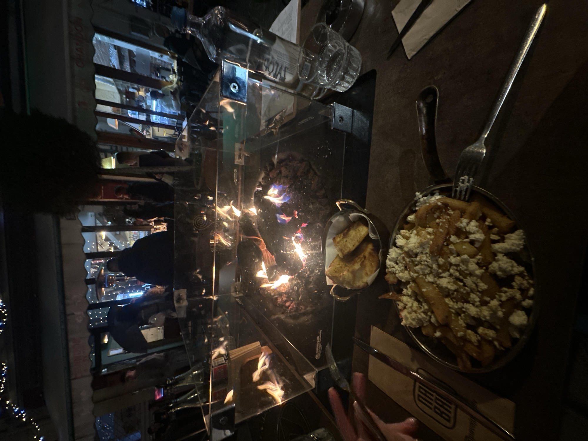 The image depicts a nighttime outdoor dining scene on a street in Plovdiv, Bulgaria, specifically at or near an establishment identified by a "PAVED" logo visible on a napkin, likely referring to Paved Bar. The immediate foreground features a dark table surface. On the table, a black skillet-style bowl contains French fries generously topped with crumbled white cheese. A silver fork rests beside it. To the left, a brown paper napkin and a knife are present, partially obscuring a person's left hand with manicured fingernails. To the right of the skillet, a small metal bowl with handles holds several pieces of toasted bread or croutons. Further right, a clear glass bottle, partially obscured, shows text suggesting "SYLVA" or similar, next to a clear glass tumbler containing liquid and ice, and an ashtray.
Central to the midground is a square-shaped, modern outdoor fire pit with a clear glass enclosure, containing active flames emanating from dark rocks, indicating a gas-fueled heat source.
The background reveals a dimly lit street scene with a building facade featuring large glass windows and an awning displaying the word "CHANDON," suggesting a related establishment or brand. The interior of this building is warmly lit, and several blurry human figures are visible inside and outside, some appearing to be patrons. Overhead, string lights with small blue bulbs illuminate the scene, contributing to the ambient lighting. Empty bottles are visible on a surface to the far left.