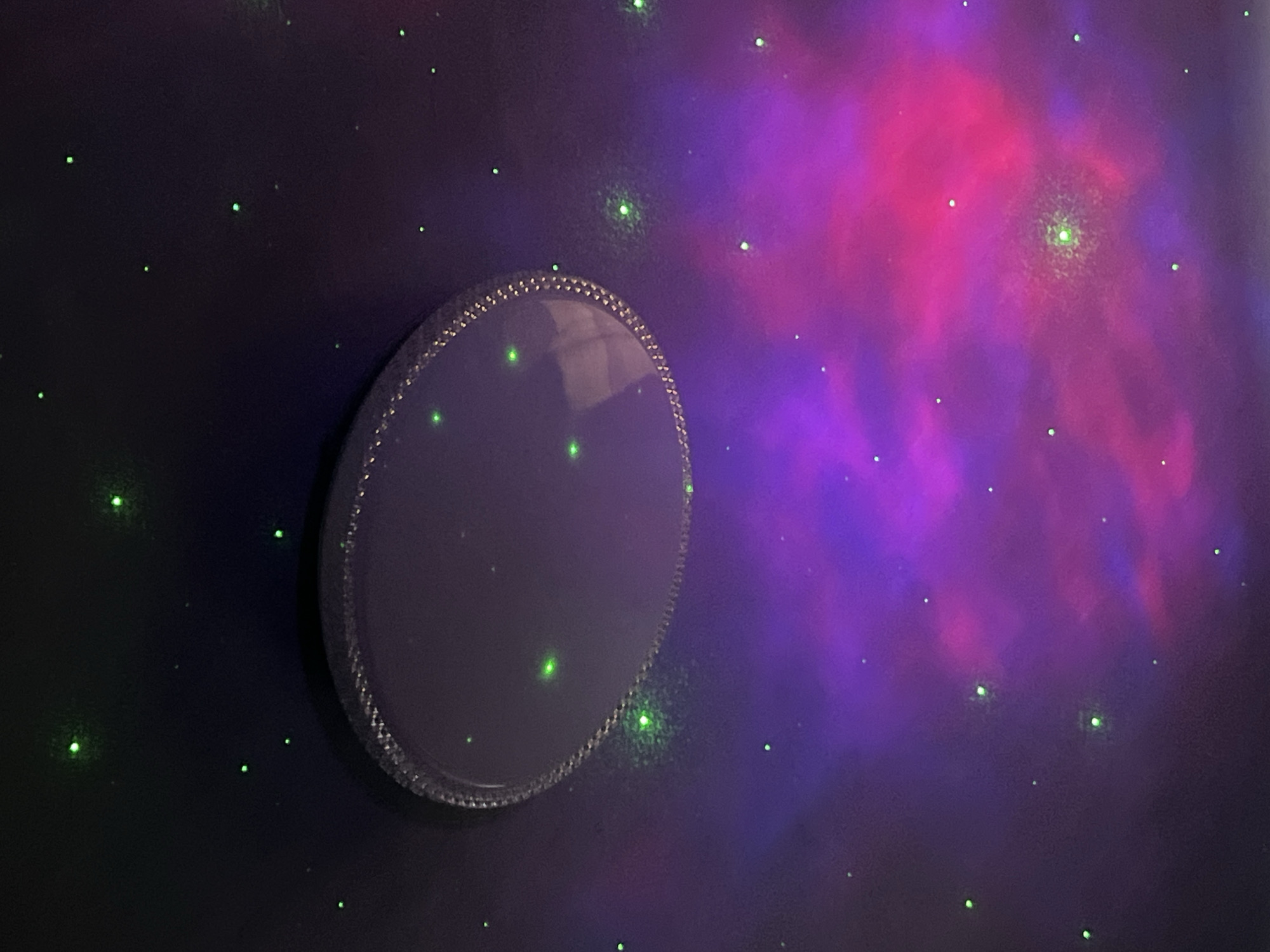 The image shows a dark room with a galaxy projection on the wall. The projection includes a nebula with pink and purple hues, and numerous small green dots representing stars. A round, possibly decorative, object with a beaded or textured edge is mounted on the wall to the left of the nebula. There is no indication of the location being in Nizhny Tagil, Russia, or any other specific city. No people, actions, or interactions are visible.