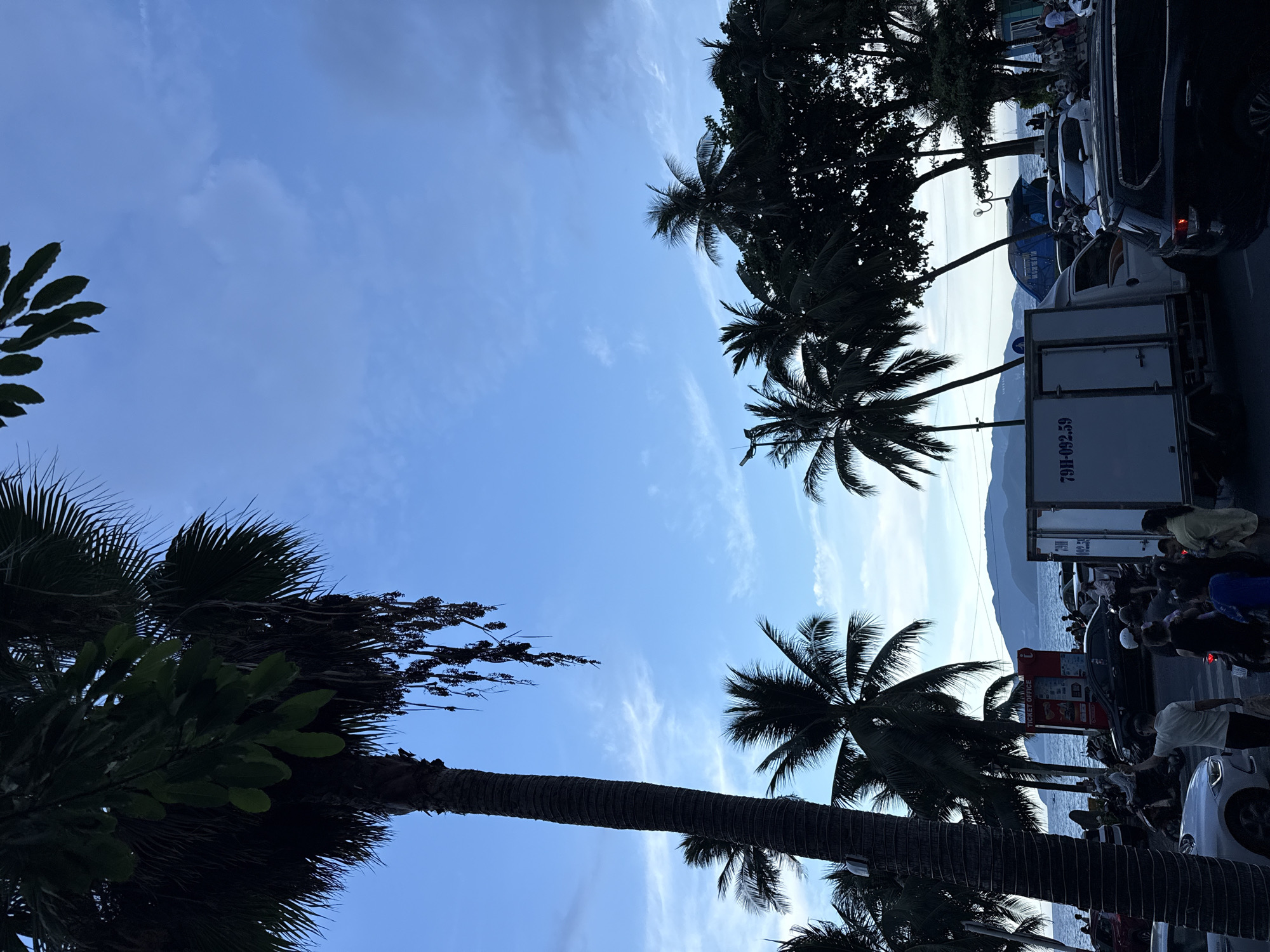 In Nha Trang, Vietnam, a street scene unfolds under a blue sky with scattered clouds and silhouetted palm trees. The foreground features a tall, textured palm tree trunk. To the right, a dark SUV is parked, its rear end visible. Further to the right, a white box truck with license plate "79H-092.59" is positioned on the road, alongside other vehicles and a blue structure. A red ticket office booth stands near the water, which is visible in the background along with distant mountains. The street is populated with numerous people, many on motorbikes, and some walking, suggesting a busy urban environment. The overall impression is of a coastal city street during daylight.