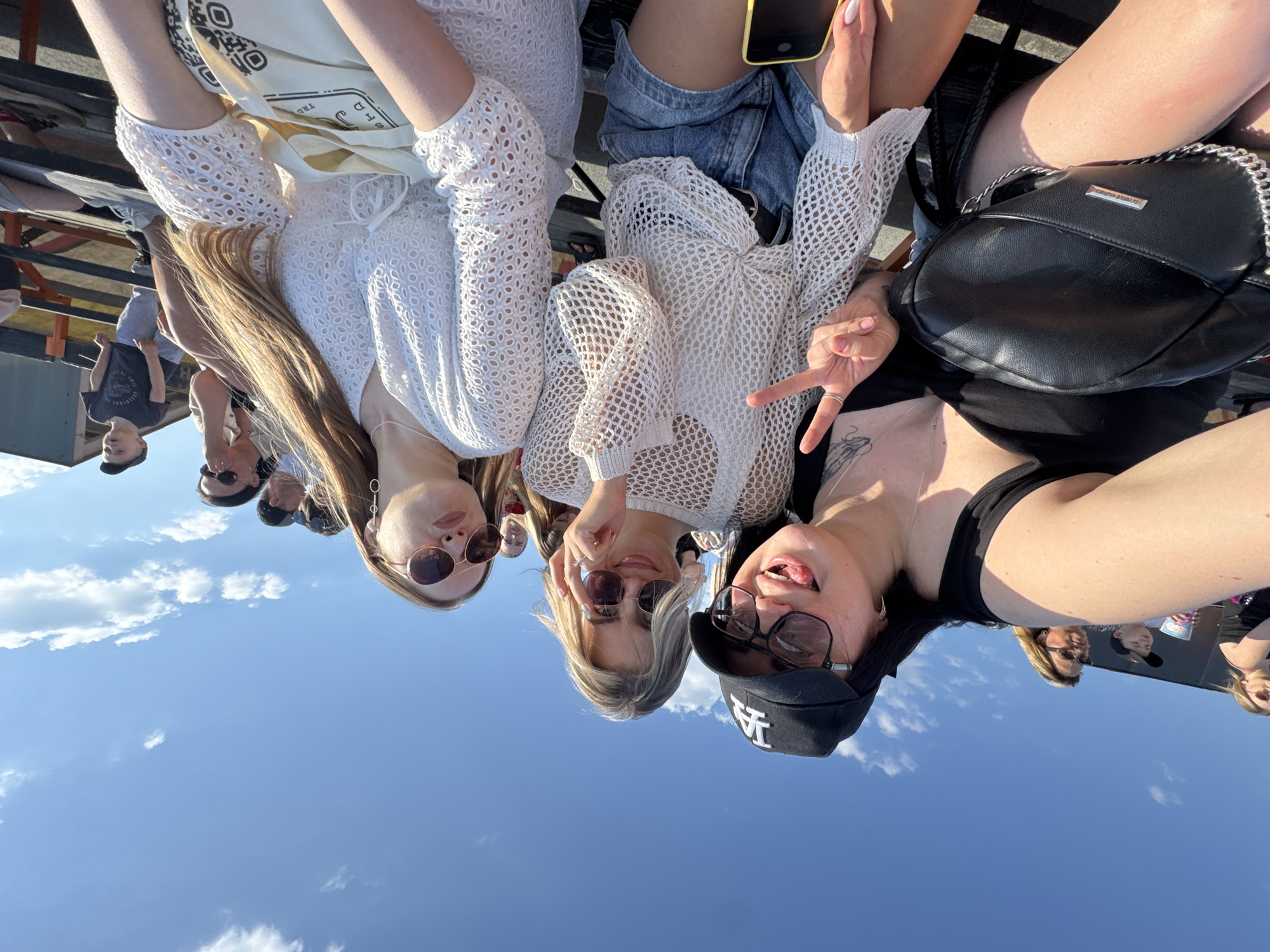 Three young women are posing for a photograph. They are in an outdoor location with a clear blue sky and light clouds in the background. They wear sunglasses and casual summer clothing. In the blurred background, multiple people are present. The location appears to be an event or gathering in the city of Unknown, Unknown.