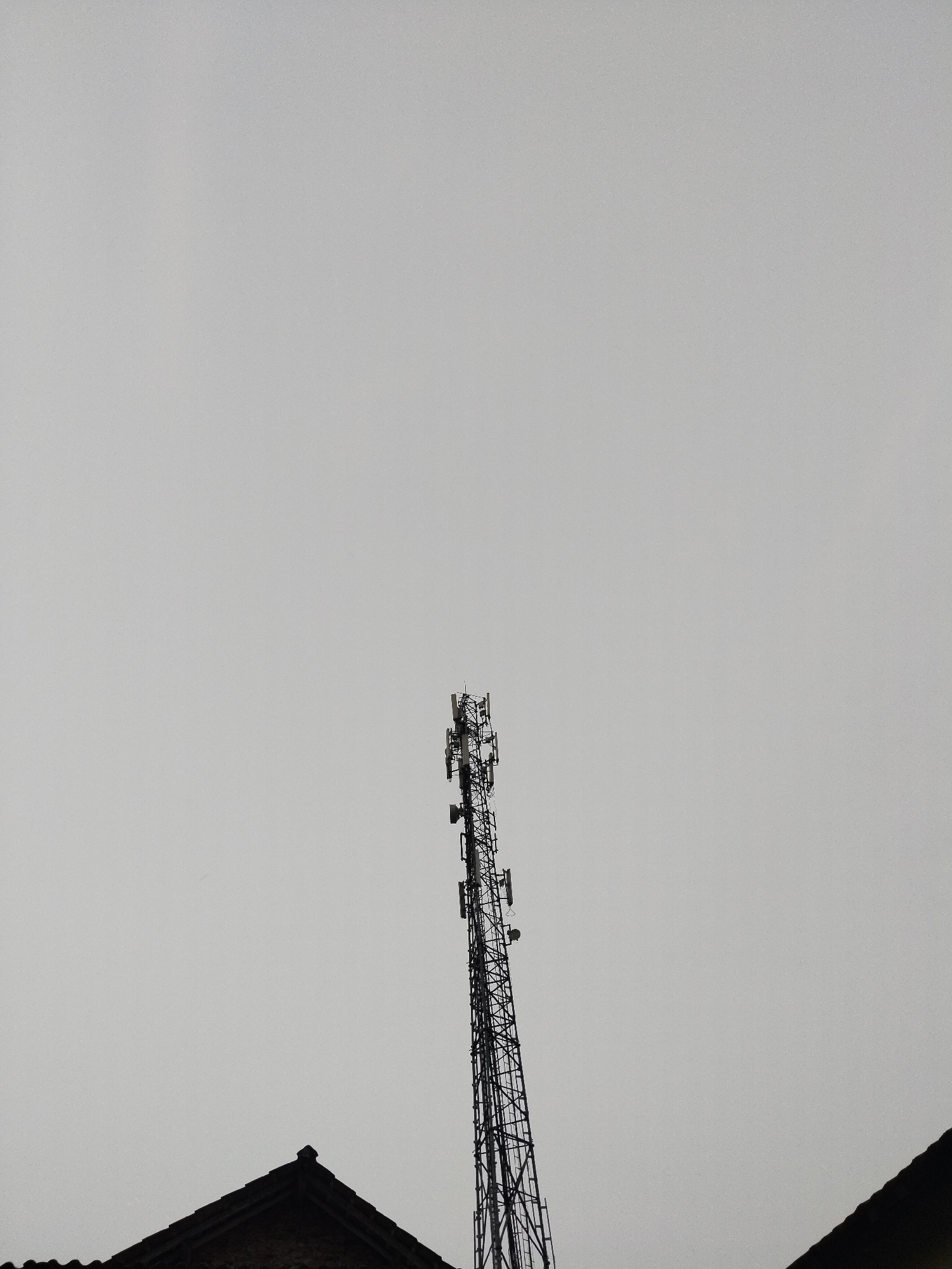 A communication tower stands tall against a overcast sky, dominating the upper-middle portion of the frame. The tower is a lattice structure, appearing dark and metallic, with multiple antennas and equipment mounted at various levels. In the foreground, the dark, silhouetted roofs of two buildings are visible, one on the left and one on the right, with their pointed gables creating a triangular shape against the lighter sky. The overall atmosphere is stark and minimalist, with the focus being on the imposing structure of the tower and the muted tones of the sky and rooftops. No people or animals are visible in the scene. The location is Kampung Baru, Indonesia.
