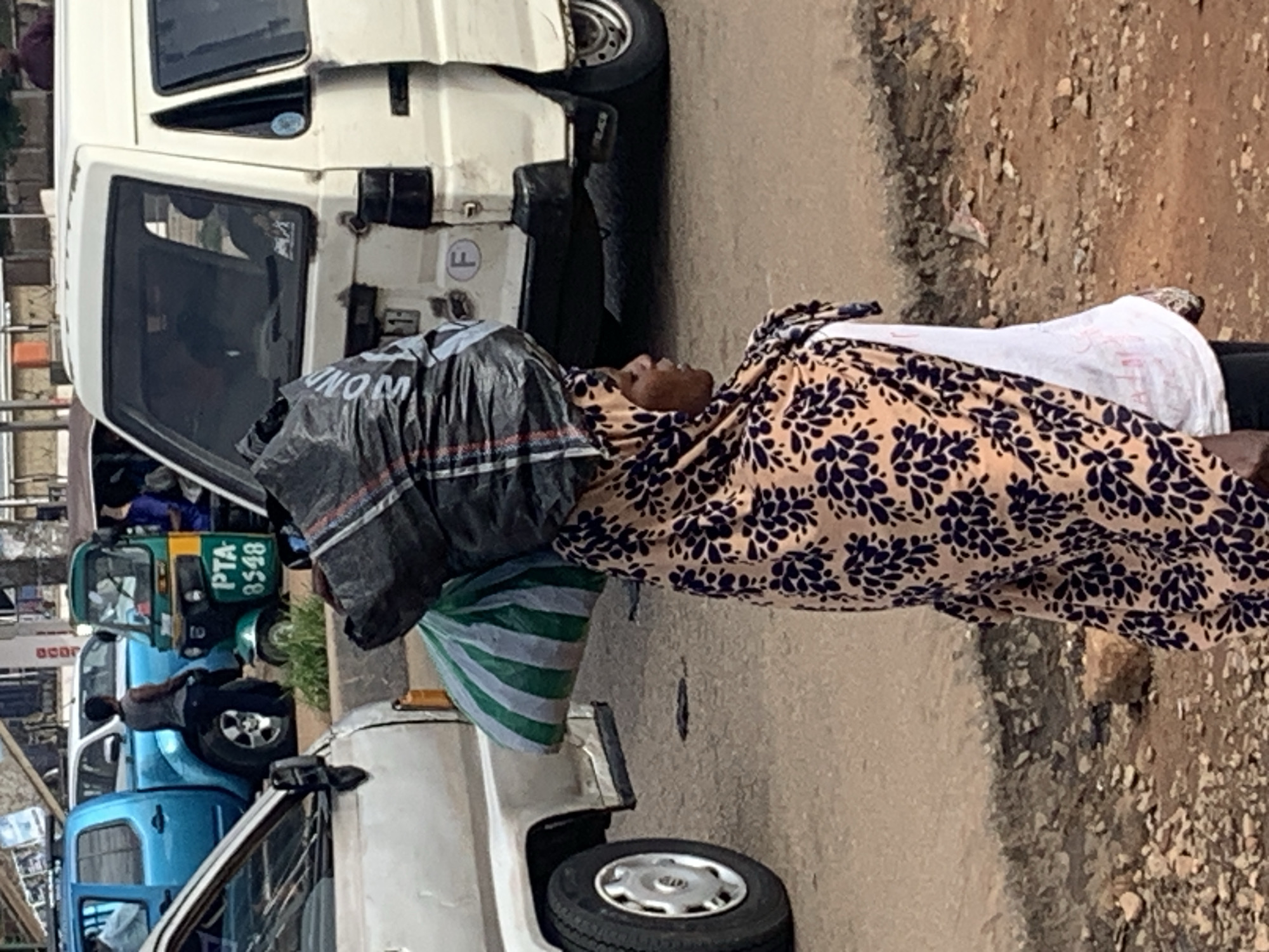In Gura Top, Nigeria, a woman is walking on a paved road with a bundle of bags on her head. She is wearing a colorful headscarf and a white shirt. Behind her, a white van and a blue pickup truck are parked. A green and yellow tricycle is also visible in the background. The road appears to be in disrepair with gravel and debris scattered around.