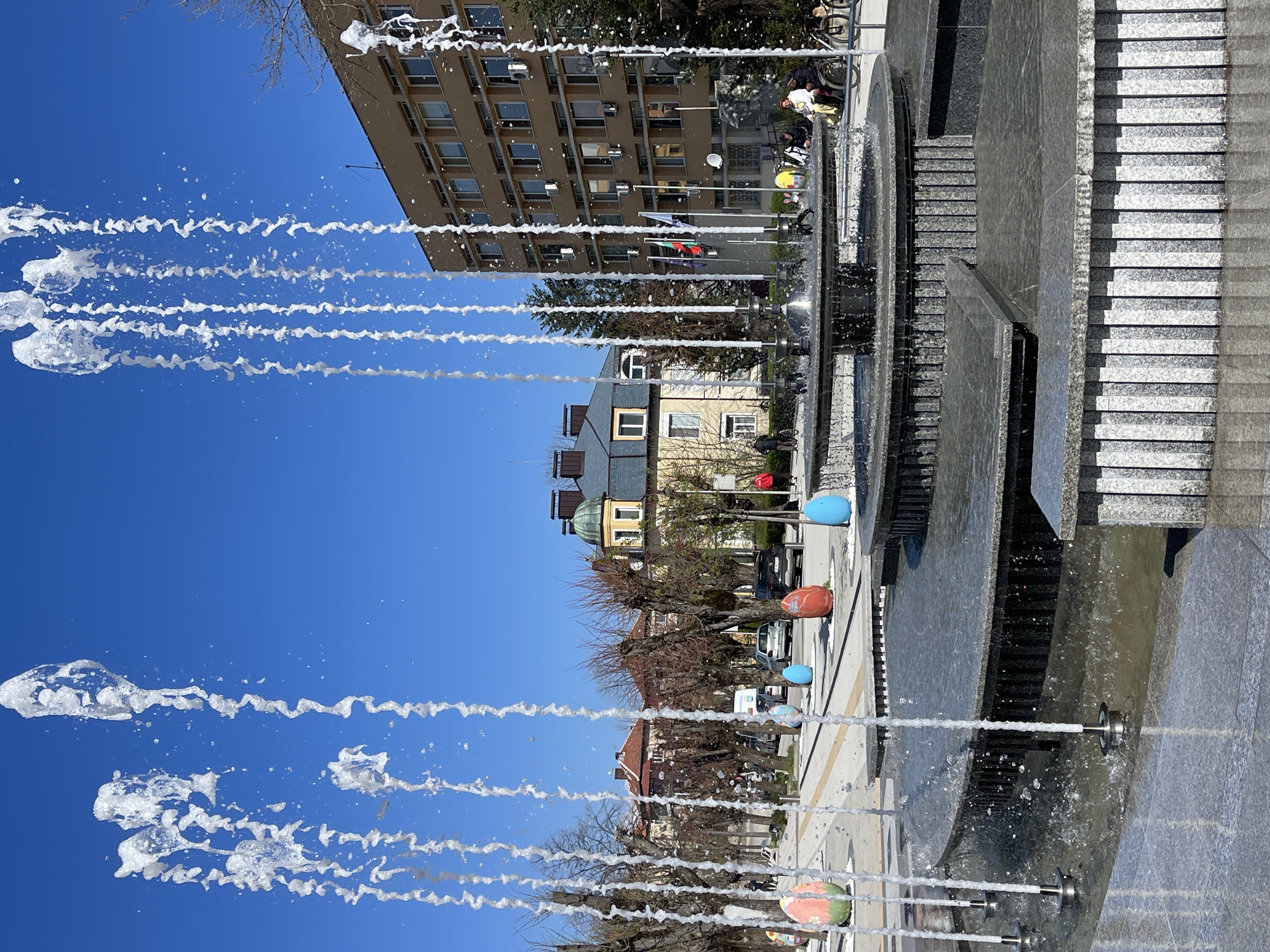 A fountain with multiple jets of water is situated in Varshets, Bulgaria, with buildings and trees in the background. The fountain is multi-tiered with stone steps and cascading water. Large, colorful decorative balls are placed along the edge. People are present near the fountain. The sky is clear blue.