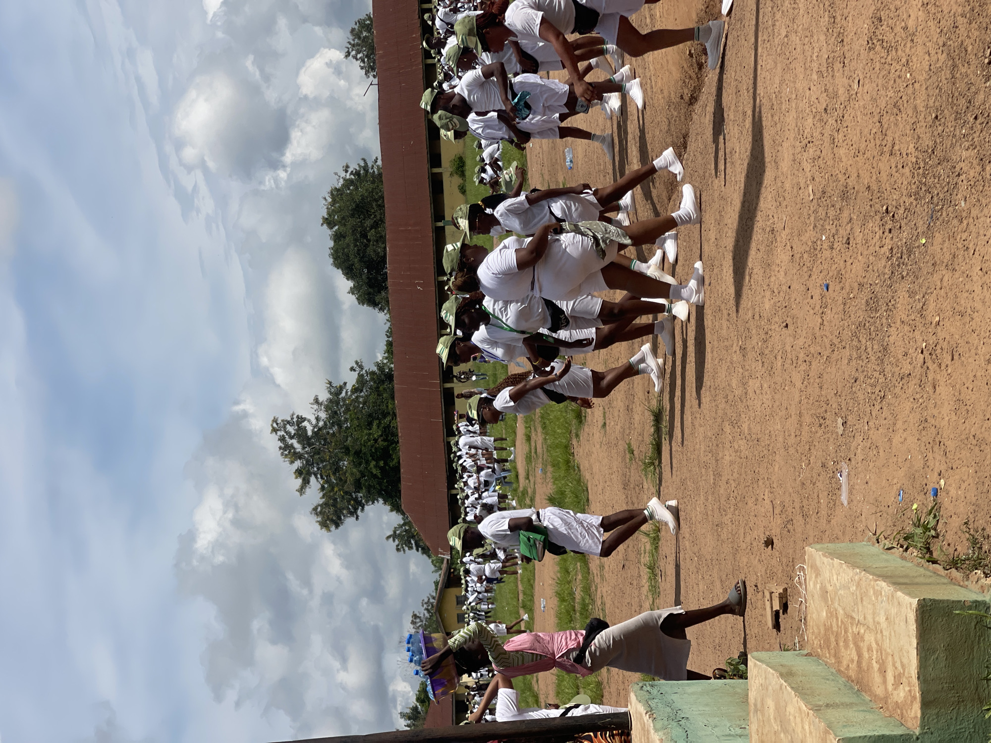In Orun Ekiti, Nigeria, a group of individuals in white uniforms and green caps are gathered outdoors on a dirt ground. Some are walking, while others appear to be standing or in formation. In the foreground, a woman with a vibrant pink and green garment walks with a basket of water bottles balanced on her head. The background features buildings with red roofs and trees under a cloudy sky.