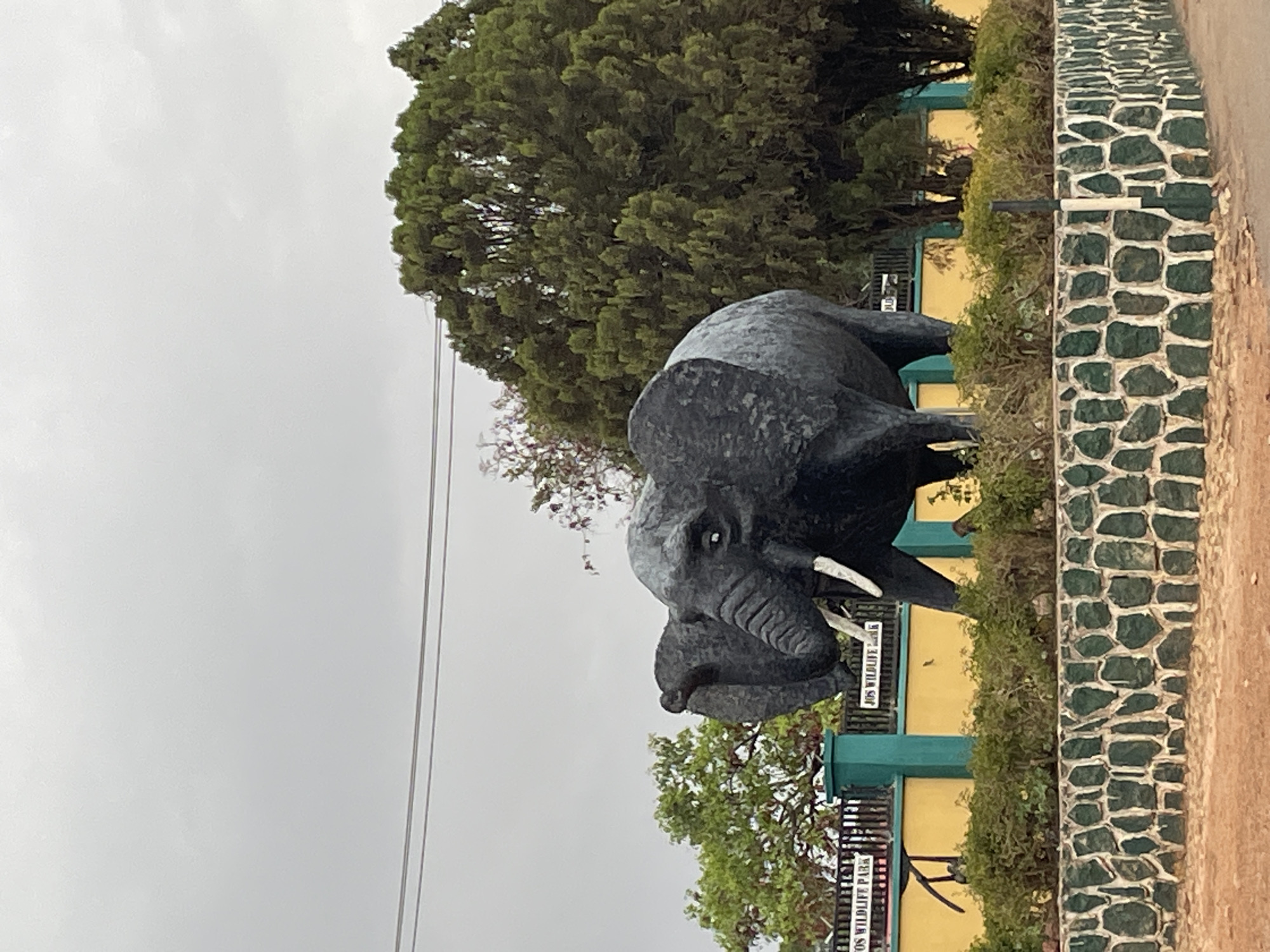 The image depicts a statue of an elephant in front of Jos Wildlife Park in Jos, Nigeria. The statue is dark grey and positioned on a raised platform with green and yellow structures behind it. A sign that reads "Jos Wildlife Park" is visible. The background includes a large green tree and a cloudy sky. A stone wall with green detailing borders the right side of the frame.