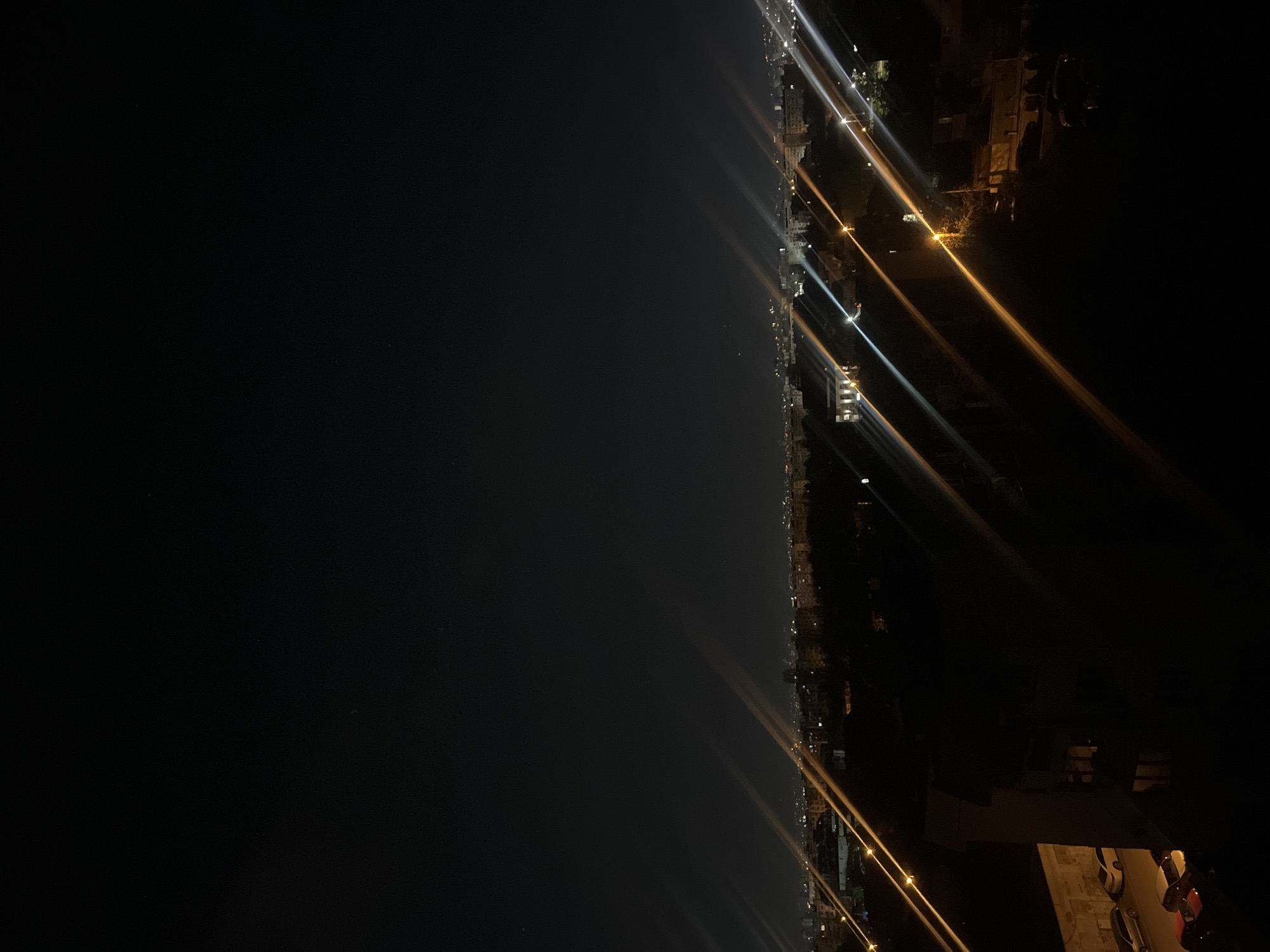 The image shows a dark scene at night, likely an aerial view or taken from a high vantage point. There is a body of water on the left side, possibly a sea or large lake, as indicated by the dark, uniform expanse. On the right side, a cityscape is visible with streetlights, building lights, and vehicle lights forming bright streaks due to the low light conditions and possible camera settings. The density of the lights suggests a coastal urban area, potentially in Sofia, Bulgaria. The buildings vary in height and are illuminated by a mix of yellow and white lights. There is no specific indication of people, actions or interactions. There are a few cars in the lower right corner.