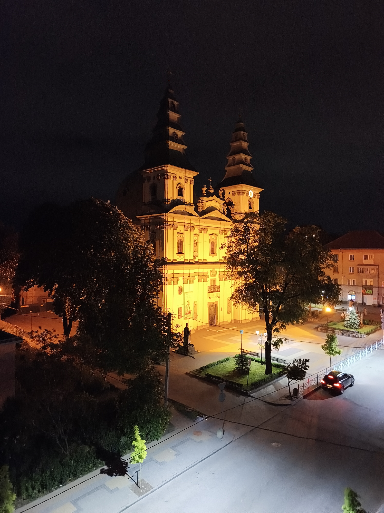 The image depicts the Cathedral of the Immaculate Conception in Ternopil, Ukraine, at night. The cathedral is brightly illuminated, showcasing its architectural details. The surrounding square is also illuminated, revealing a few trees and landscaping. A dark-colored car is parked on the street near the square. The sky is dark and clear. The lighting suggests a well-lit urban environment.