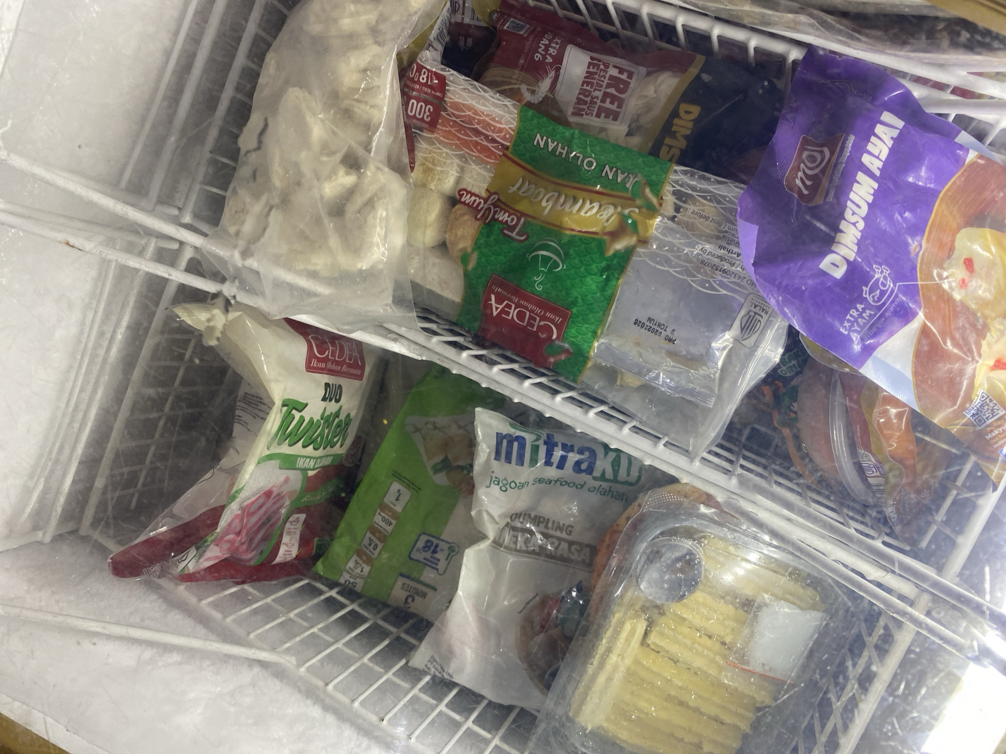 This image depicts the interior of a chest freezer filled with various frozen food items. The freezer is divided into compartments by wire racks. Visible food items include bags of dim sum, frozen dumplings, and what appears to be sliced cheese or butter in a plastic container. Some packaging is labeled in Indonesian, with brands like "Mitraku," "CEDEA," and "Dimsum Ayai." The environment is cold and icy, as evidenced by the frost on the freezer walls. The image was taken in the city of Payatieng, Indonesia.