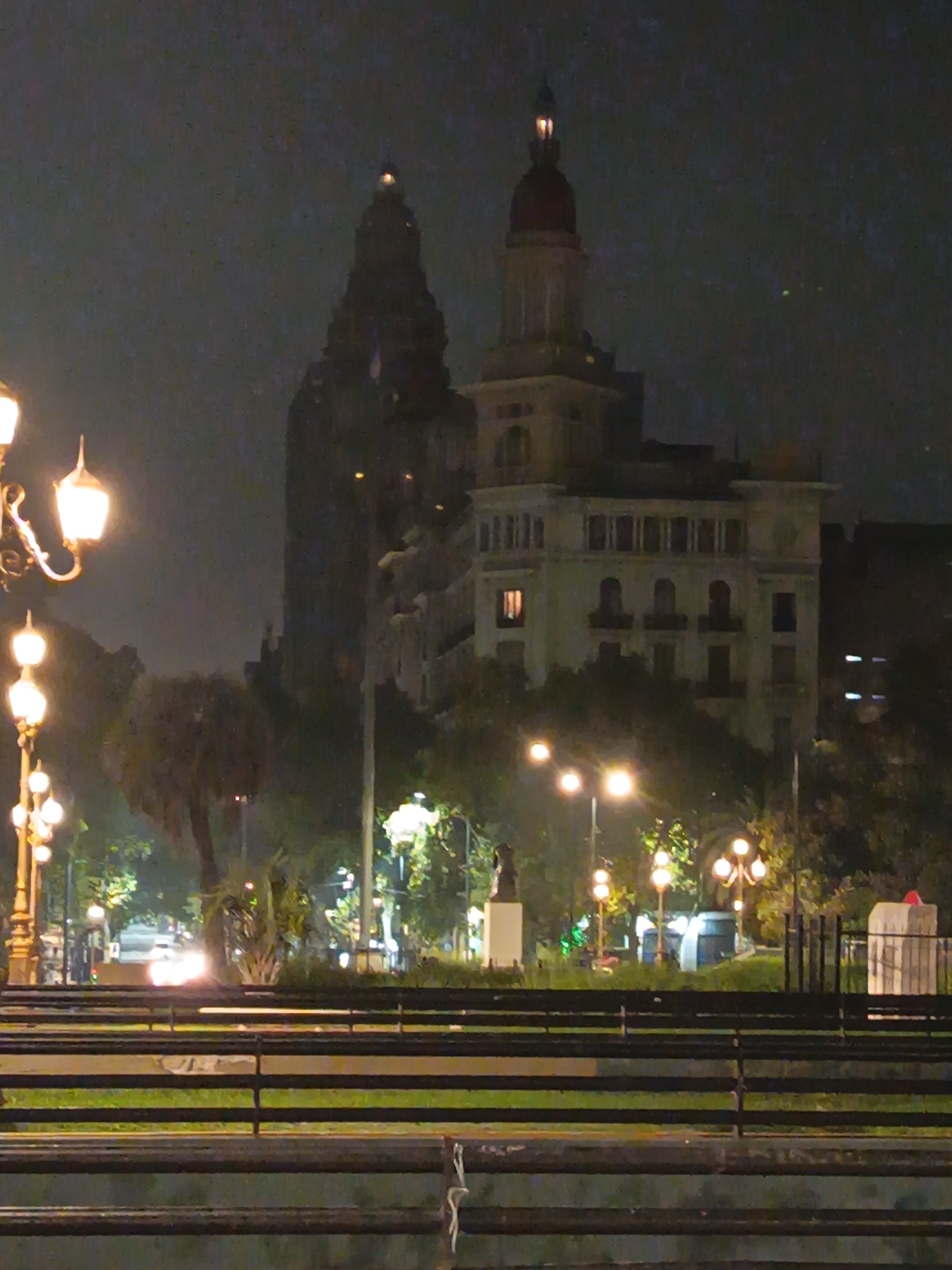 This nighttime image depicts a cityscape in Buenos Aires, Argentina, focusing on the illuminated Palacio Barolo and the Edificio de la Prensa. A line of ornate street lamps illuminates the foreground. A park or plaza is visible in the midground. Benches and a low fence are in the foreground. No people are distinctly visible. The sky is dark and overcast.