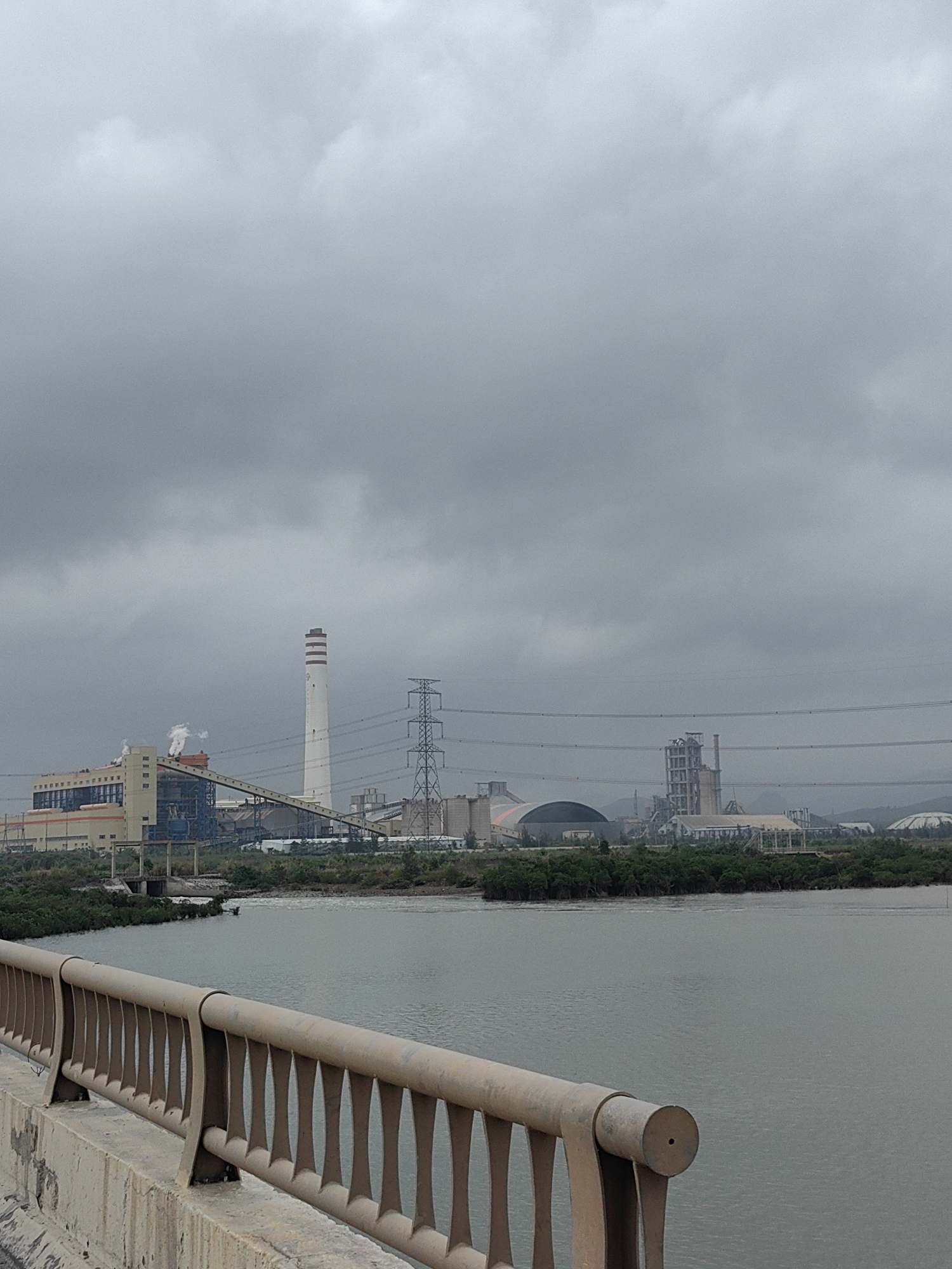 The image presents an expansive industrial landscape under a heavily overcast sky, viewed from a bridge. In the immediate foreground, a robust concrete bridge railing with decorative, light-colored metal spindles runs along the bottom of the frame.
The midground is dominated by a wide, calm body of water, likely a river or estuary, which separates the viewer from the distant industrial complex. The far bank of the water is lined with dense, dark green vegetation, possibly mangroves or other riverside plants.
In the background, a large industrial facility spans the horizon. On the left, a cluster of buildings with light beige and blue exteriors is visible, some emitting plumes of white smoke or steam from their stacks, indicating active operations. A long, dark conveyor belt system extends diagonally across this section. Central to the industrial scene is a very tall, slender white chimney, marked with three horizontal dark bands near its apex. To its right, a tall metal electricity transmission tower stands, supporting multiple power lines that stretch across the frame. Further right, more industrial structures can be seen, including large dome-shaped storage facilities and a tall, multi-level concrete building, possibly part of a cement or processing plant. Faint outlines of low hills or mountains are visible in the distant background.
The sky is uniformly covered with thick, gray clouds, suggesting a completely overcast day, possibly humid or preceding rain. No specific human activity or visible text is depicted. The scene captures an industrial zone located in Thong Nhat Commune, Vietnam.