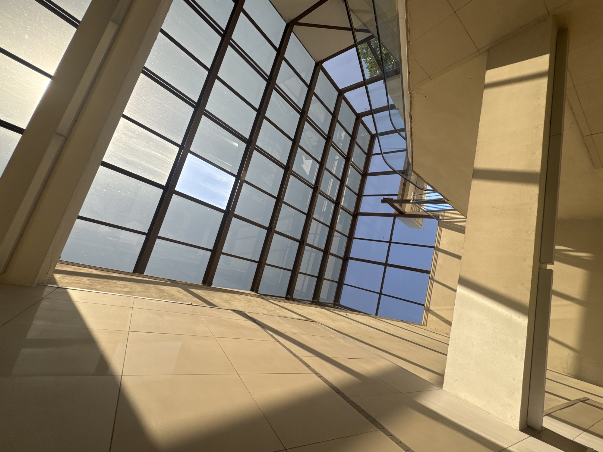 The image depicts the interior of a modern building with a multi-pane glass roof. Sunlight streams through the glass, casting long shadows on the beige-colored walls and structural elements. The scene is an atrium or similar open space, with a staircase and what appears to be an escalator railing visible in the background. The architectural style suggests a public or commercial building. The lighting suggests it is daytime. There are no people visible in the frame. The location is identified as Sofia, Bulgaria.