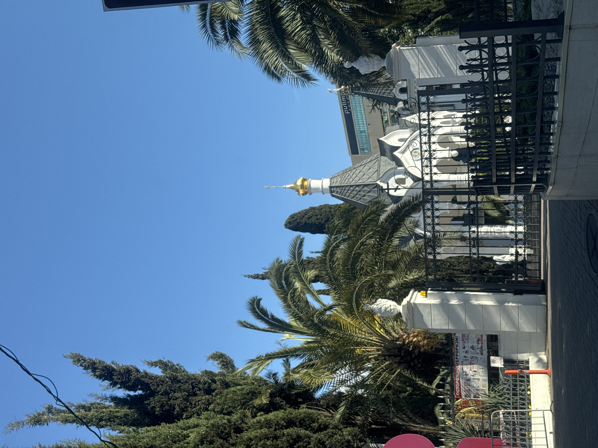 The image captures a view of the Chapel of St. Nicholas the Wonderworker in Sochi, Russia, situated near the Sochi Pullman Hotel. The chapel, with its golden dome, is framed by palm trees and other greenery against a clear blue sky. A black iron fence surrounds the chapel grounds, and a sidewalk runs alongside it. The building of Sochi Pullman Hotel visible behind the chapel. There is also a sign written in Russian at the base of the church.