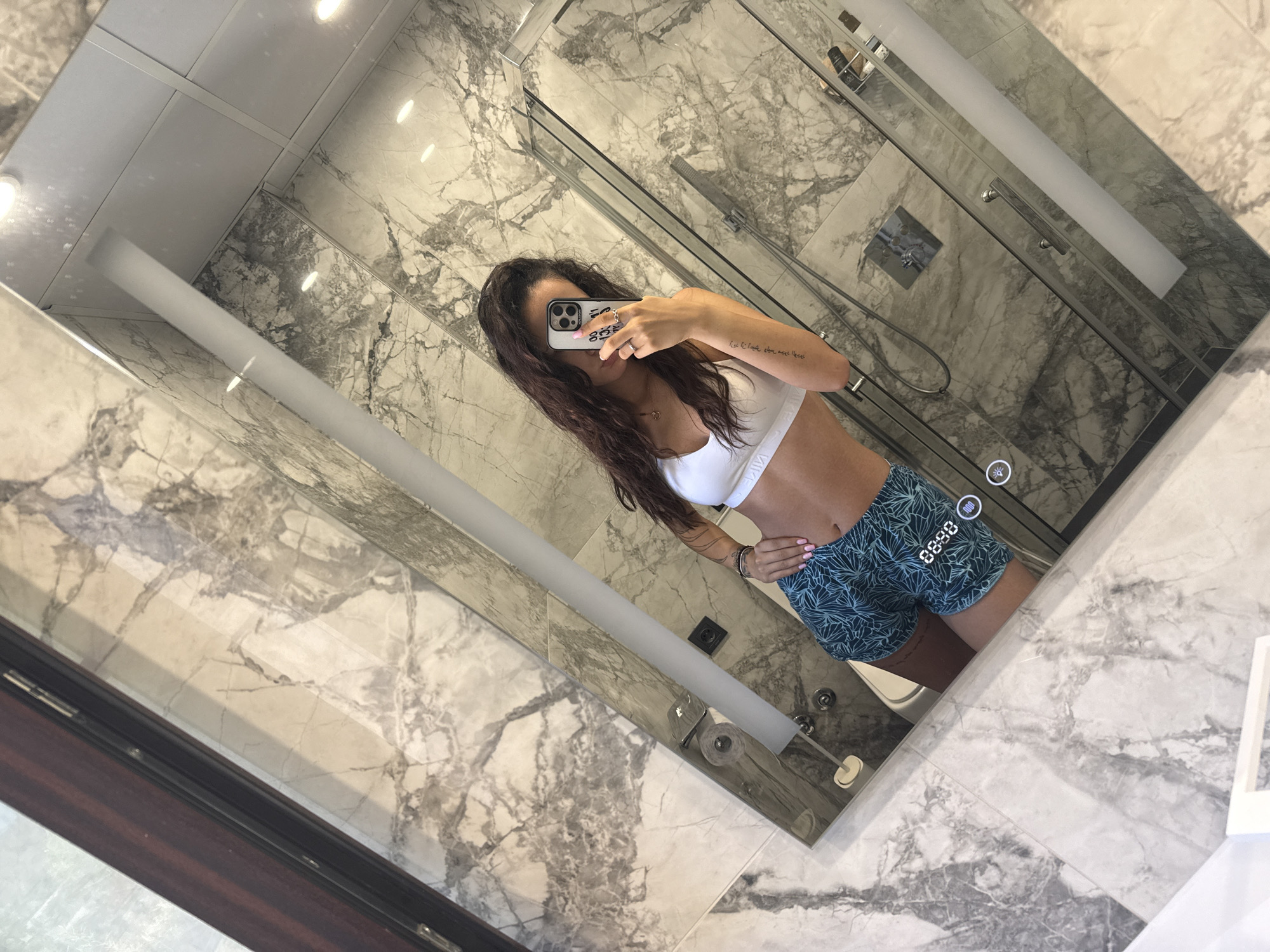 A young woman with long, dark, wavy hair is taking a selfie in a modern bathroom. She is wearing a white sports bra and teal patterned shorts. Her body is angled, and she is holding a smartphone in her right hand, obscuring her face. She has tattoos on her left arm and is wearing rings on her fingers. The bathroom features marble walls and a glass shower enclosure. A toilet is visible behind her. The lighting in the bathroom is bright and reflective. The image captures a section of a vanity mirror that reflects the woman and the bathroom surroundings. The overall scene suggests a casual, private moment.