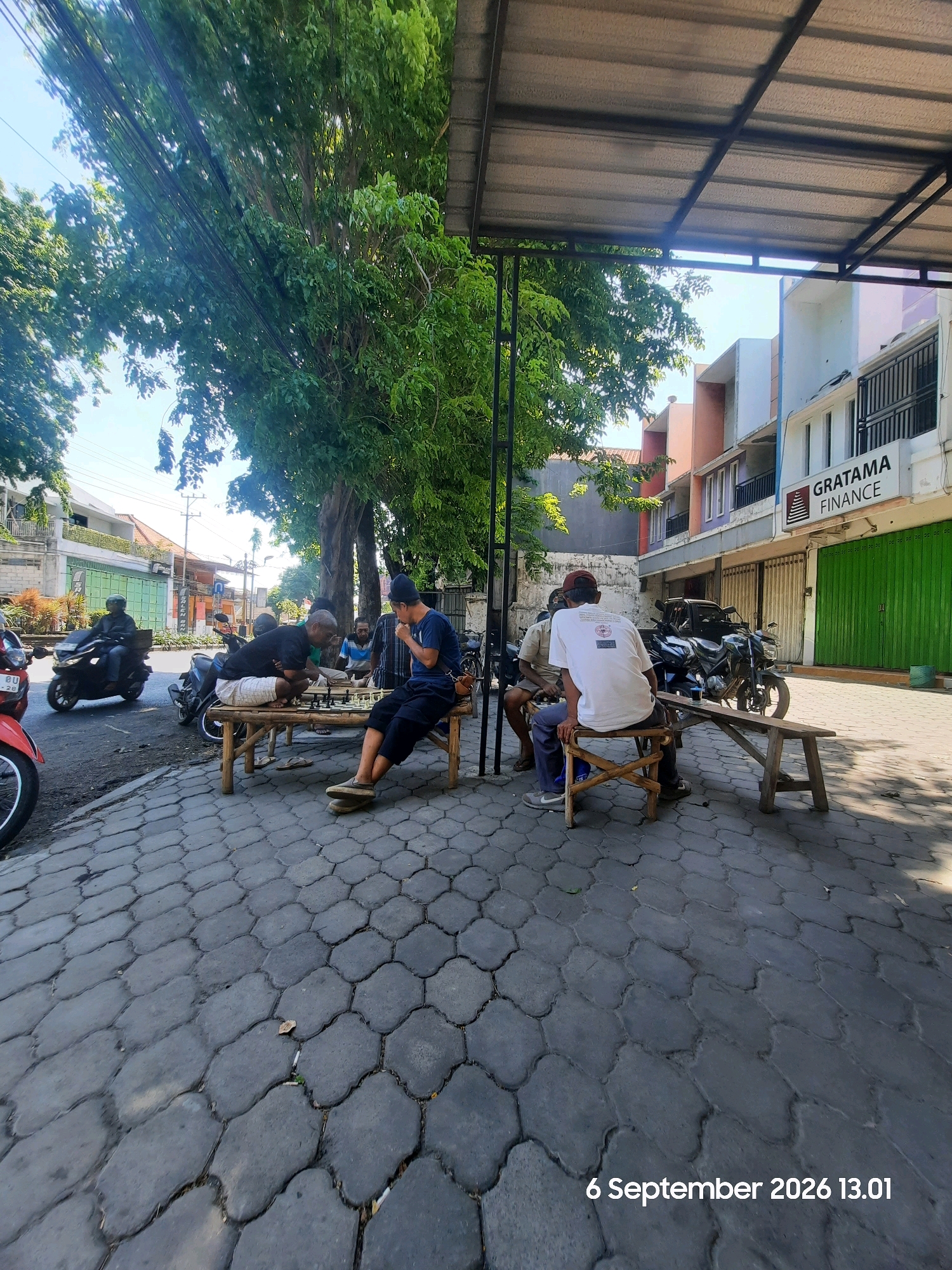 In Probolinggo, Indonesia, a group of men are playing chess outdoors on a paved sidewalk. The scene is captured on September 6, 2026, at 13:01. They are gathered around a wooden table under the shade of a large tree, with a makeshift awning providing additional cover. A few motorcycles are parked nearby, and in the background, buildings with shop fronts, including one labeled "GRATAMA FINANCE," line the street. One of the men, wearing a blue shirt and black pants, is in the foreground, seemingly contemplating a move. Other individuals are seated or standing around the chess board, engaged in the game. The ground is paved with dark, interlocking hexagonal tiles, and the overall atmosphere suggests a casual, communal activity taking place on a sunny day.