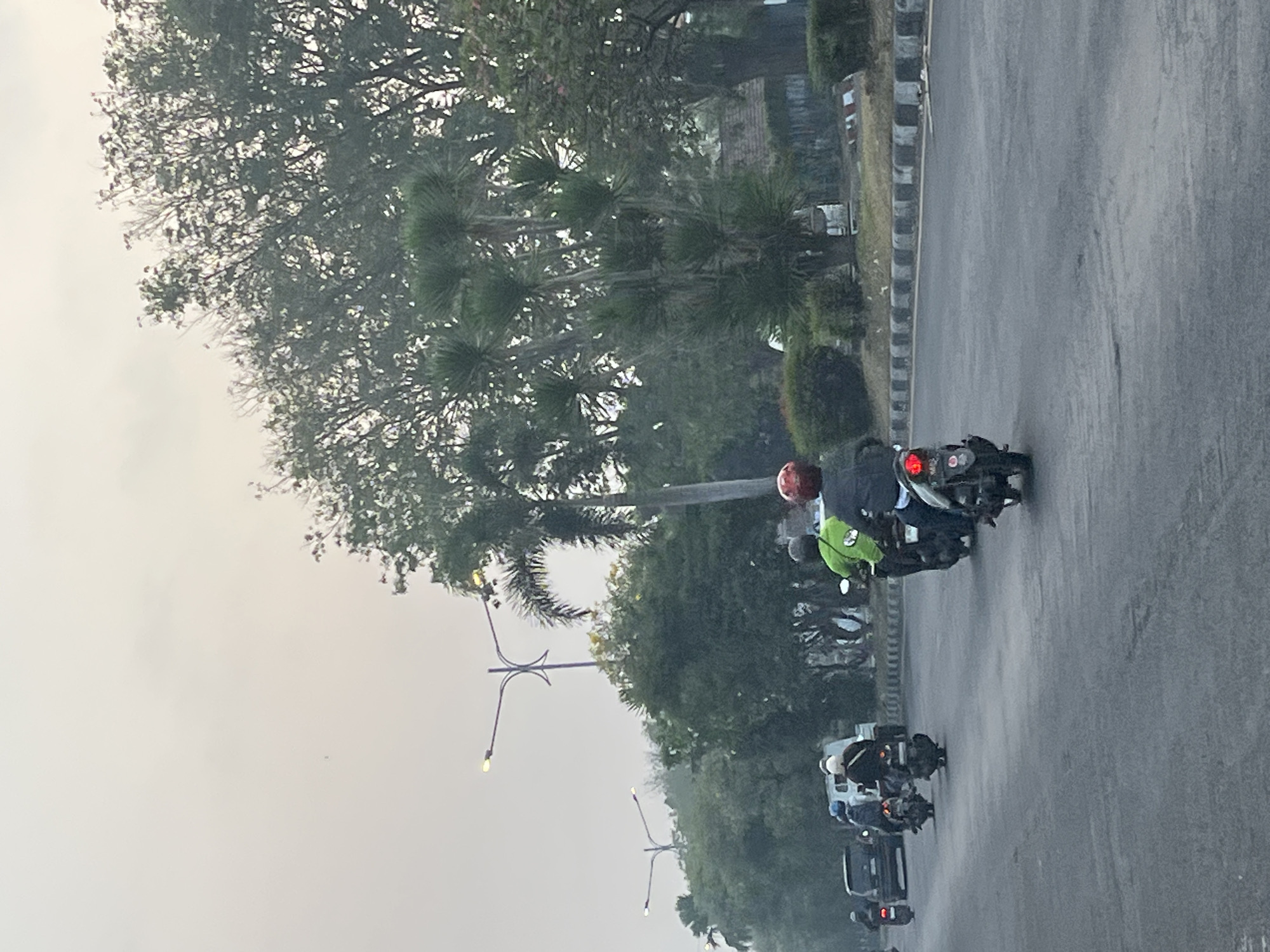 The image captures a street scene in Kludan, Indonesia, under an overcast sky. Several motorcycles are traveling on a paved road. On the right side of the road, three individuals are riding motorcycles. The furthest motorcycle has a rider wearing a green shirt and a dark helmet. The motorcycle in front of that has a rider wearing a red helmet. On the left side of the road, several other motorcycles are visible, along with a white van. The road is bordered by a curb and a grassy area with trees and bushes. Tall palm trees are also present in the background, and streetlights are visible along the road. The overall atmosphere appears to be calm, with traffic moving at a moderate pace.