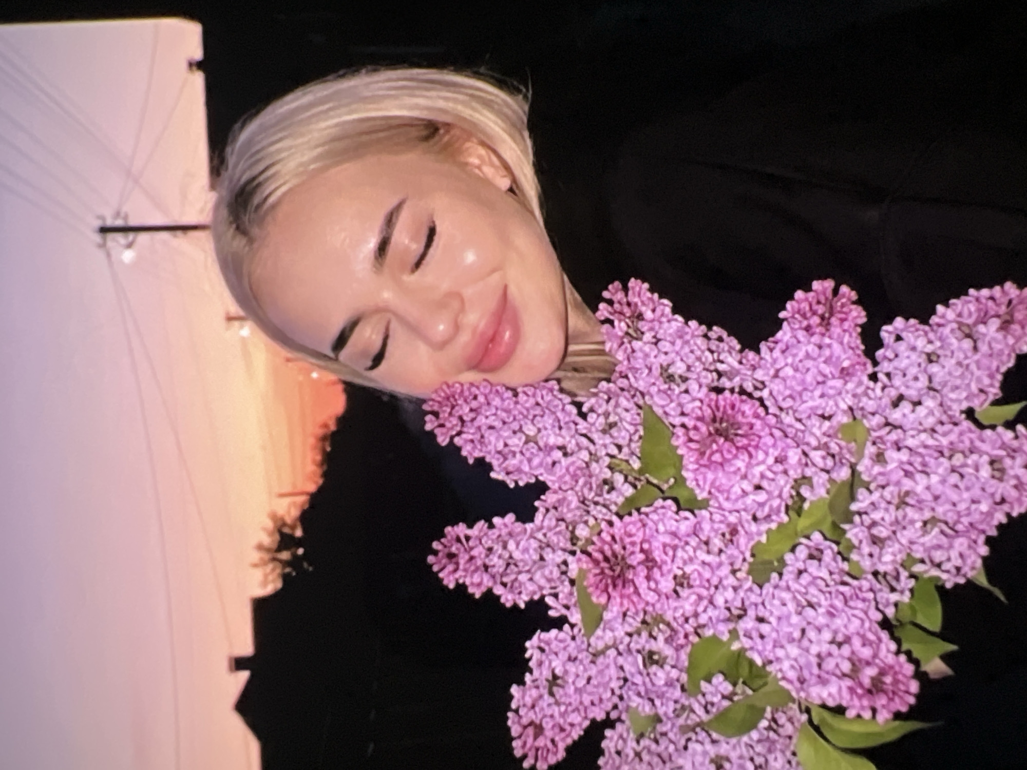 A young woman with blonde hair and fair skin is shown with her eyes closed and a serene, slightly smiling expression, resting her head against a large bouquet of vibrant purple lilac flowers. Her head is tilted, and she appears to be enjoying the presence of the flowers. She is wearing dark clothing that blends into the shadowed right side of the background.
The bouquet of lilacs is a prominent feature, displaying clusters of small, densely packed purple and pink blooms with a few green leaves visible. The background is divided: the right side is predominantly dark, suggesting night or deep shadow, while the left side features a soft, pastel-colored sky. This sky transitions from shades of light pink and orange near the horizon to a paler lavender-blue higher up, indicating either a sunrise or sunset. Faint, thin dark lines, resembling utility wires or power lines, are visible against the lighter sky, and a dark silhouette of a distant building or structure with a peaked roof can be seen near the horizon. The overall ambiance of the image is calm and peaceful. No text is visible in the media file.