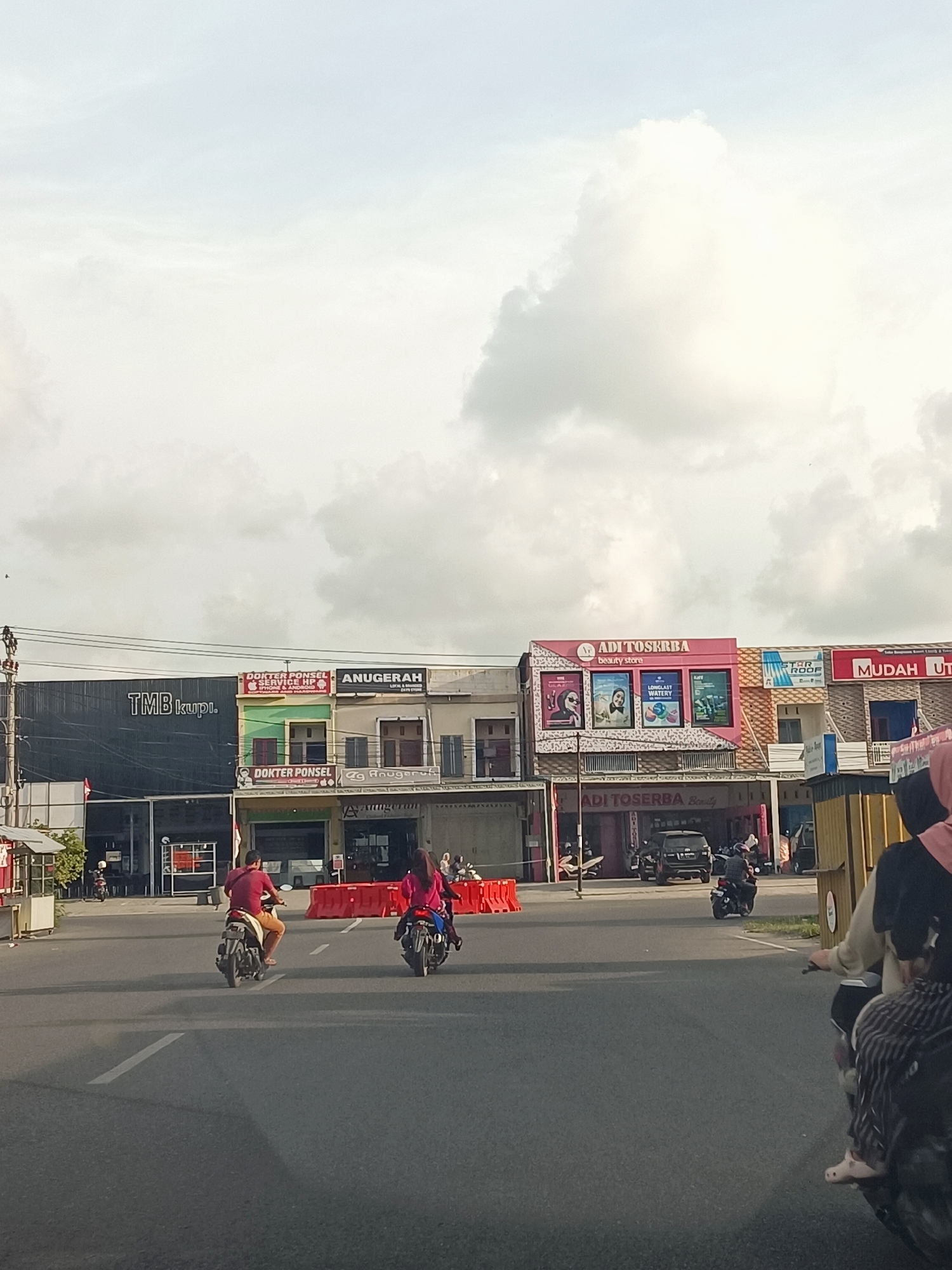 The image captures a street scene in Sigli, Indonesia. In the foreground, an asphalt road with lane markings is visible. Two motorcycles are traveling on the road, one with a male rider wearing a red shirt and tan shorts, and another with a female rider wearing a pink hijab and jacket. In the background, a row of shops and buildings lines the street. One building displays a prominent sign that reads "TMB kupi.". Other signs indicate businesses like "DOKTER PONSEL SERVICE HP" and "ANUGERAH beauty store". Several orange traffic barriers are placed along the sidewalk. The sky above is cloudy with bright patches. A dark-colored SUV is parked in front of one of the buildings. The overall environment suggests a commercial area within the city.