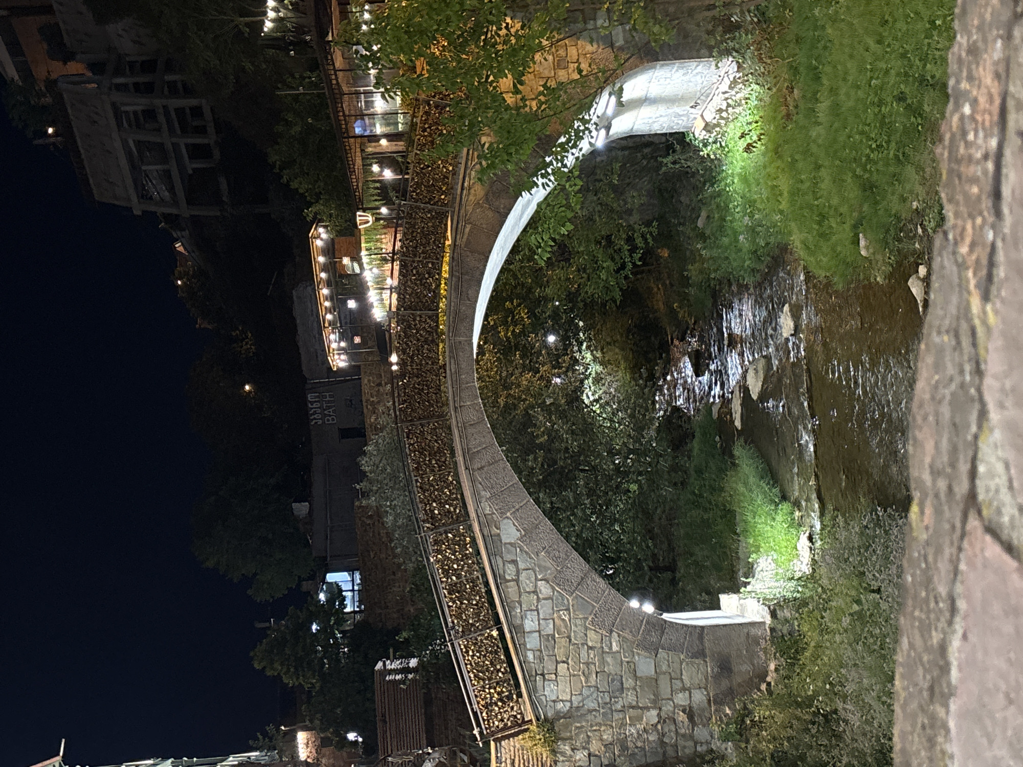 An arched stone bridge at night, covered in padlocks on its railing, spans a shallow stream. The bridge is illuminated by lights, and the surrounding area is overgrown with trees and foliage. In the background, buildings with lights are visible, suggesting a populated area. A dark night sky is overhead.