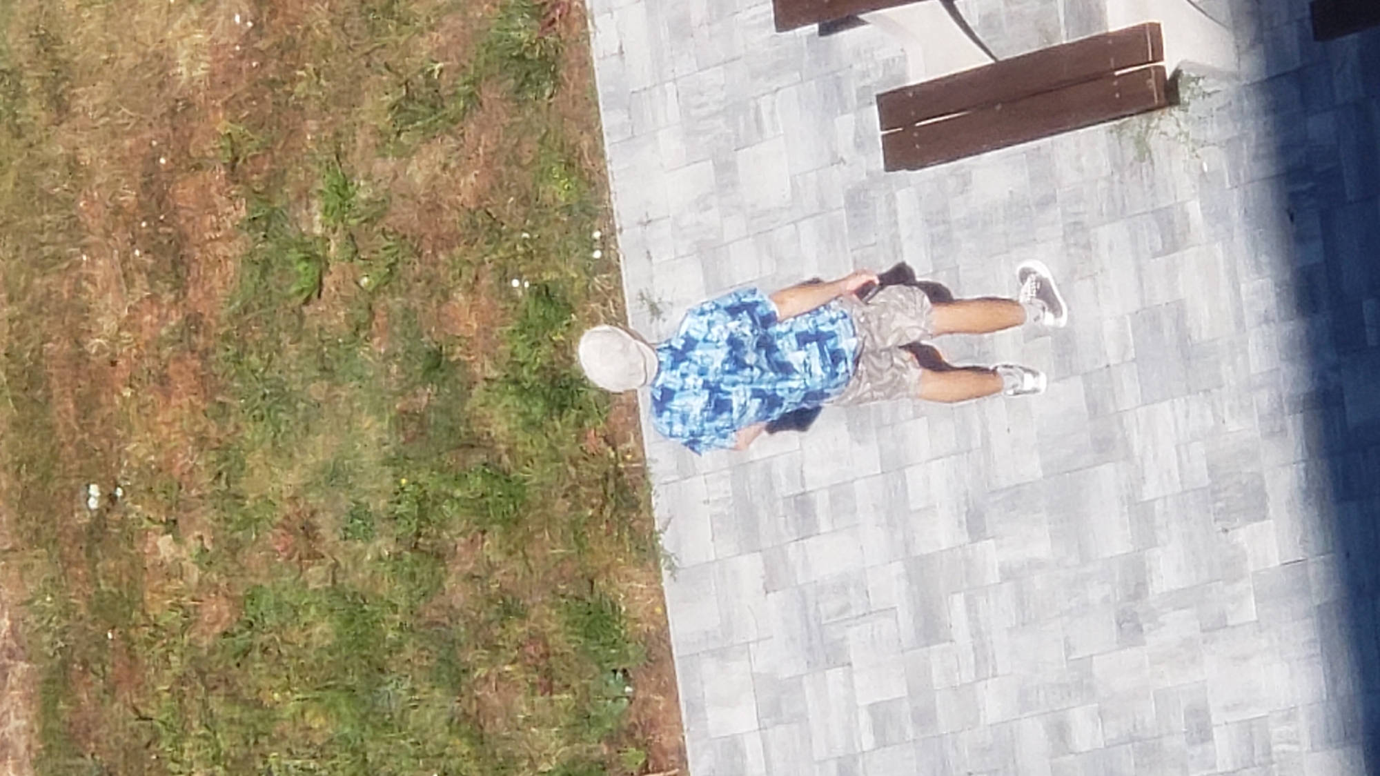 An overhead shot captures a person from behind, standing on a paved patio area. The individual is wearing a light-colored flat cap, a short-sleeved shirt with a blue and white abstract pattern, camouflage-patterned shorts, and white sneakers. The patio is made of grey rectangular pavers, and beyond it, a patch of dry, brownish-green grass is visible. A wooden bench with dark brown planks is partially visible on the right side of the frame, positioned on the patio. The scene appears to be outdoors on a sunny day. The location is not specified beyond being a city, and no specific landmarks are identifiable to determine the exact location within Prešov, Slovakia.