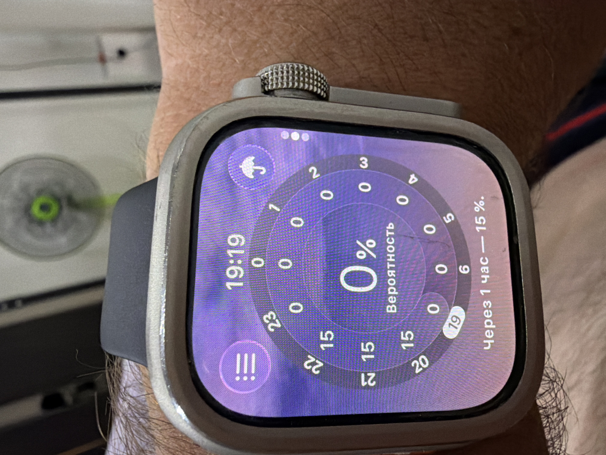The image shows a close-up of a person's wrist wearing a silver smartwatch with a dark band. The smartwatch display shows the time as 19:19 and features a circular graphic with numbers and percentages, indicating weather information. A purple icon with three horizontal lines is on the left side of the screen, and an umbrella icon is on the right. Below the circular graphic, text in Russian reads "Вероятность" (Probability) and "Через 1 час — 15 %" (In 1 hour - 15%). The background is blurred but appears to be an indoor setting with a fan visible. There are no discernible elements that would place the image in Baan Mae Nam, Thailand.