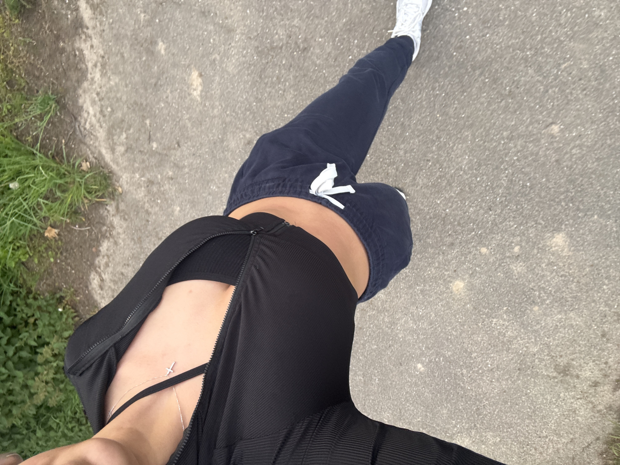 A high-angle full body shot shows a person walking on a paved surface. The person is wearing a black sports bra under a black, ribbed zip-up jacket that is partially open. The person is also wearing dark blue sweatpants with a white drawstring. One white sneaker is visible in the top of the frame. To the left is a patch of green grass along the edge of the pavement. The location cannot be determined by the image.