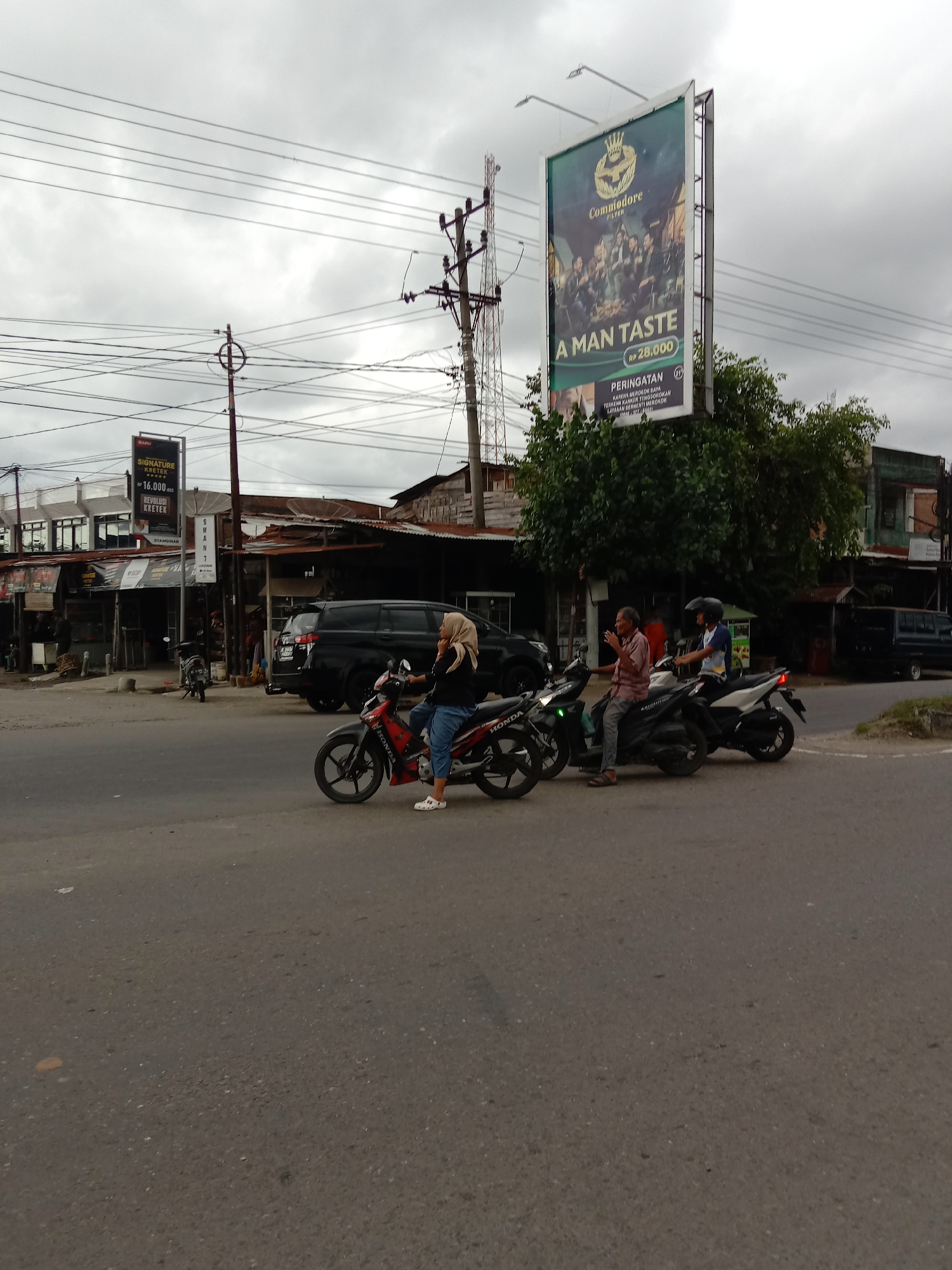 The image depicts a street scene in Lhokseumawe, Indonesia, under an overcast sky. The scene is a busy intersection with several motorcycles stopped or waiting. The image includes various signs, including those advertising "SIGNATURE KRETEK" and "Commodore Filter" cigarettes. Several people are riding or standing next to motorcycles, including a woman wearing a hijab who is on her phone. Buildings and shops line the street in the background, along with parked cars and utility poles.