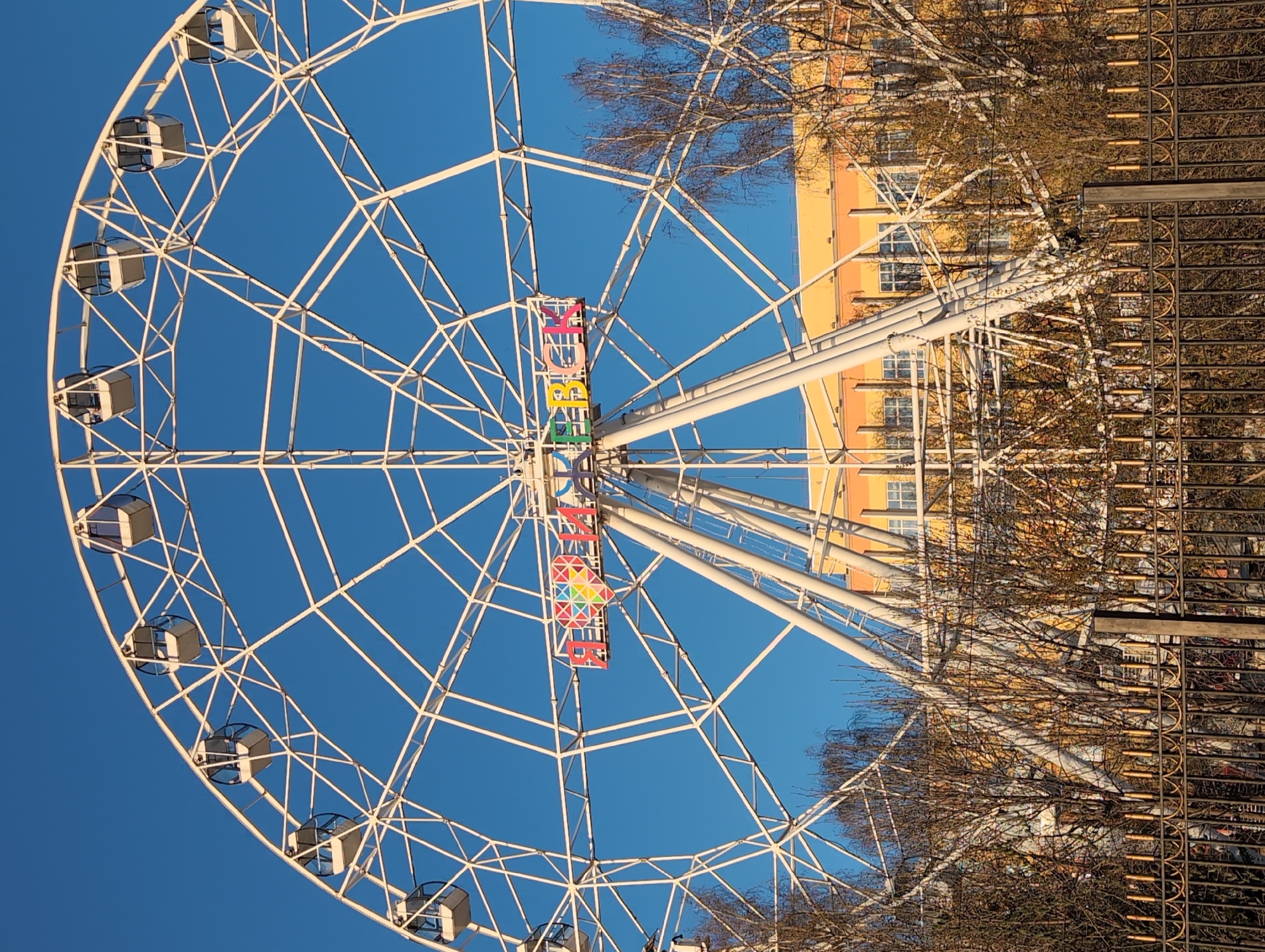 A daytime, low-angle shot features a white Ferris wheel against a clear blue sky, with the word "OBZOR" (translated as "Overview" or "Observation") displayed vertically in colorful, illuminated letters at its center. The Ferris wheel has enclosed passenger cars spaced around its circumference. In the background, a yellow building is partially visible behind bare trees and a dark metal fence. This Ferris wheel is located in the city of Izhevsk, Russia.