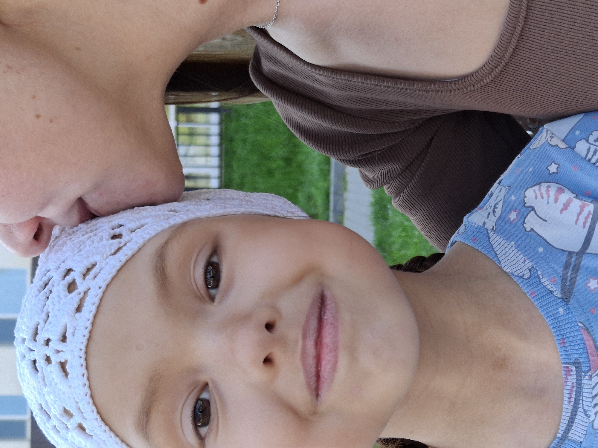 A mother kisses her child on the forehead. The child is wearing a white crocheted cap and a blue pajama top with a cat pattern. The mother is wearing a brown top. The location is Chaikovsky, Russia, as indicated by the image's metadata. The scene appears to be outdoors near a fenced area and greenery.