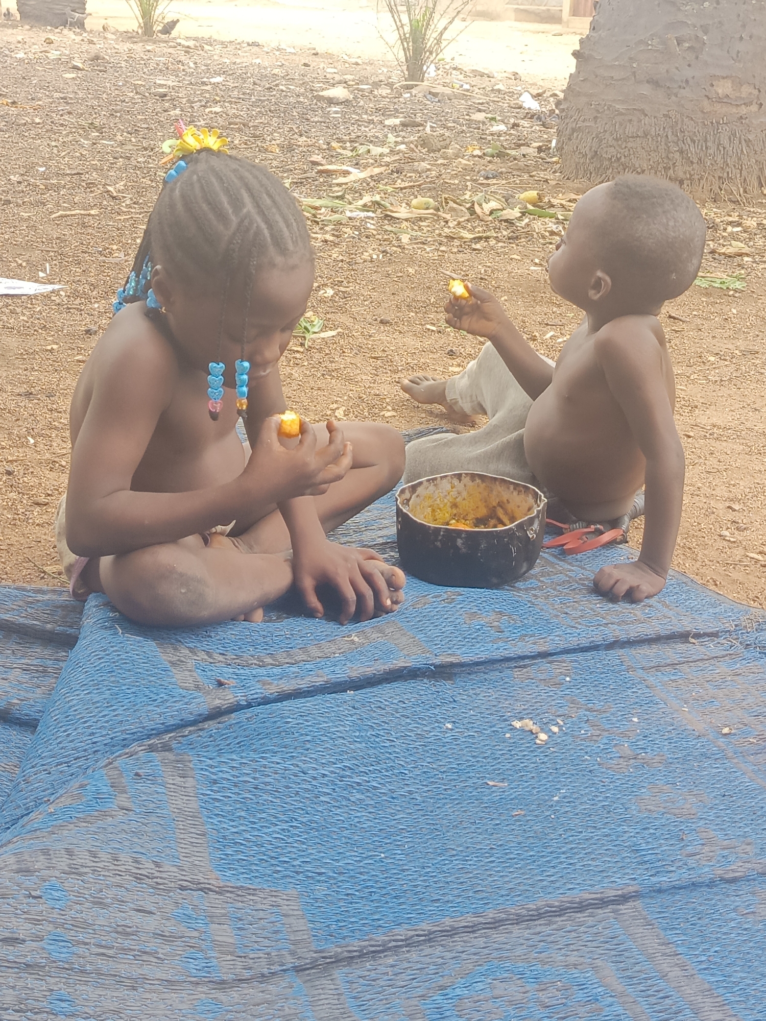 Two children, a girl and a younger boy, sit on a blue woven mat outdoors. The girl, with braided hair adorned with a yellow flower and blue beads, sits cross-legged, holding a piece of orange food. The boy, shirtless and wearing light-colored pants, also holds a piece of the same orange food. A black bowl sits between them, appearing to contain more of the orange food. The ground around the mat is dirt and scattered debris. The location appears to be Akwanga, Nigeria.