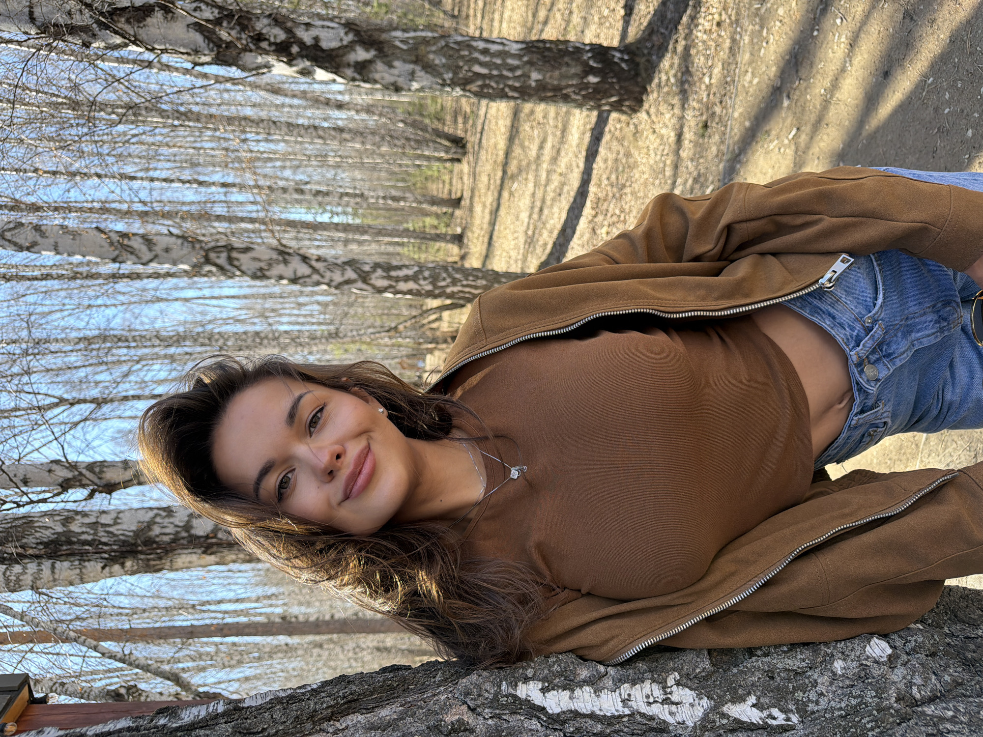 A young woman is captured in a casual outdoor portrait, leaning on the trunk of a large birch tree. She has long, dark brown hair and a pleasant smile, looking directly at the camera with her head slightly tilted. She wears a fitted brown long-sleeved top, a light brown zip-up jacket, and light blue denim jeans. A silver chain necklace with a small pendant and stud earrings are visible.
The setting is a birch forest or park, characterized by numerous tall birch trees with their distinctive white and dark-patterned bark. Many of the trees are bare of leaves, suggesting either early spring before foliage emerges or late autumn after leaves have fallen. A clear blue sky is visible through the tree branches in the background. The ground is covered with dry leaves and dirt, with patches of bright sunlight and distinct shadows cast by the trees, indicating a sunny day. The scene evokes a relaxed and natural atmosphere. The abundance of birch trees aligns with the provided location context of Krasnoyarsk, Russia. No visible text is present.