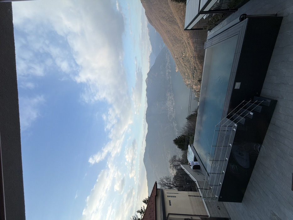 The scene depicts an elevated outdoor pool area in Brunate, Italy, offering a panoramic view of Lake Como and the surrounding mountainous landscape. A dark overhead structure partially frames the immediate foreground. A large, rectangular infinity-edge swimming pool with blue water and a dark-toned base occupies the center-lower portion of the image. Metal stairs with handrails provide access to the pool on its left side. Adjacent to the pool on the left, a hot tub is situated on a raised platform. One individual, clad in a white robe, stands with their back largely to the camera, positioned next to the hot tub. A multi-story building with a red-tiled roof is partially visible on the far left. A dark metal fence runs along the property's edge overlooking the valley. On the right, a white-roofed pergola structure stands adjacent to the pool. The midground prominently features Lake Como, characterized by its deep blue water and visible shoreline settlements, nestled between steep, tree-covered hillsides. The background comprises multiple layers of mountains, with distant peaks exhibiting traces of snow. The sky is partially clear blue with dispersed white cumulus clouds. The overall illumination suggests late afternoon or early evening daylight.