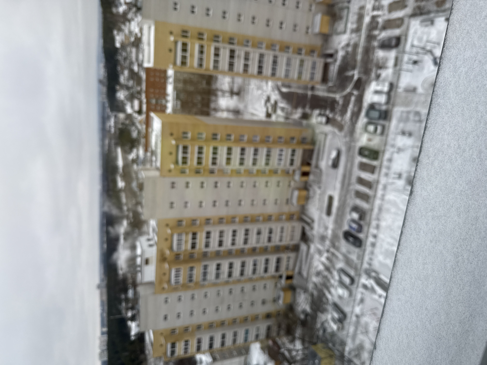 The image shows an aerial view of a residential area in Yekaterinburg, Russia, likely during winter. Several high-rise apartment buildings with a distinctive repetitive window pattern dominate the scene. The buildings appear to have a yellowish-beige color along the window lines. Between the buildings, snow-covered grounds are visible, along with parked cars and possibly some roads. The sky is overcast and the environment appears cold. The focus is not sharp, suggesting the image might be taken from a moving aircraft or drone.