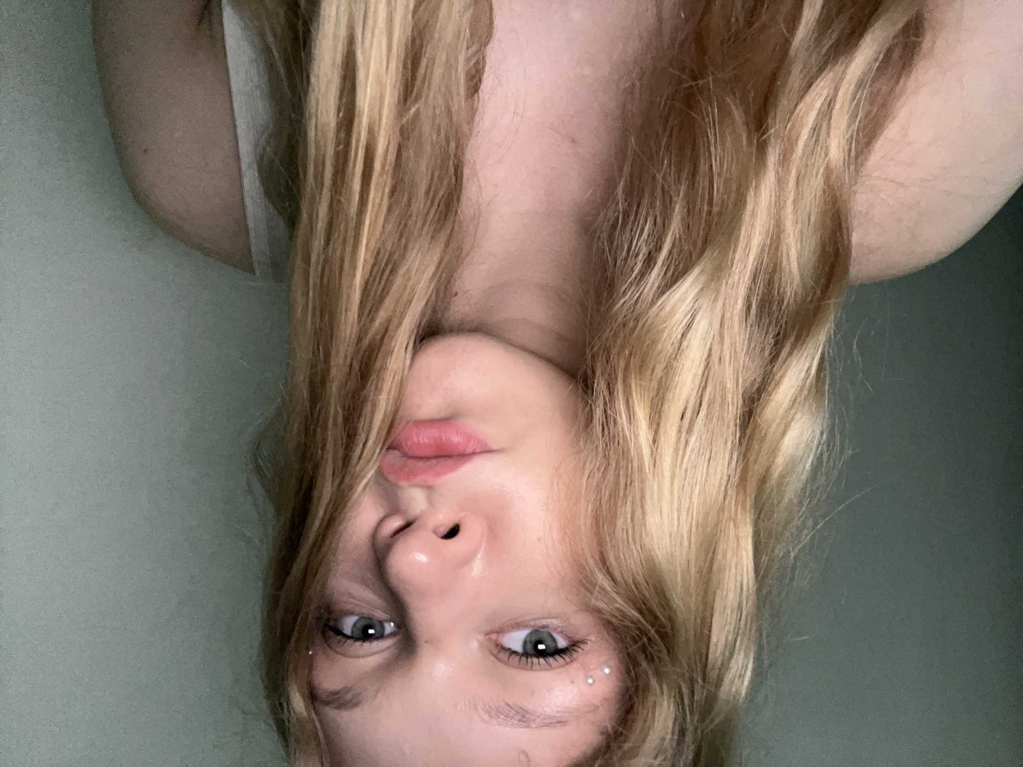 A young woman with blonde, wavy hair and light blue eyes is pictured in a close-up, front-facing shot. She is wearing a light-colored tank top. Small, shimmering studs are placed on her left eyebrow. The background is a plain, pale green wall. The scene does not provide any information to ascertain its location in the city of Perm, Russia.